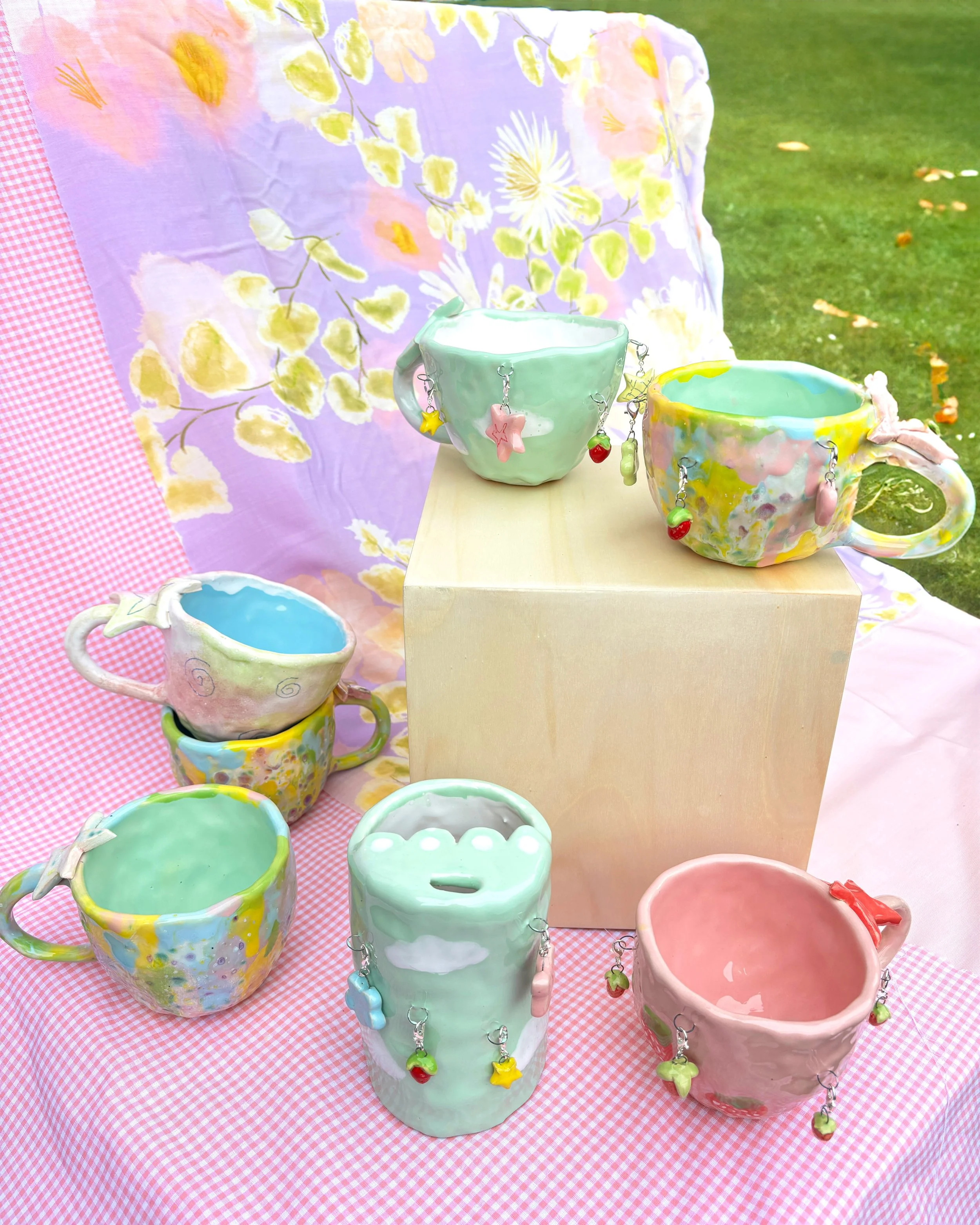 Colorful ceramic mugs with charms hanging on the handles are displayed on a wooden platform, with a pink and white checkered tablecloth underneath, and a floral patterned fabric and grass background behind.