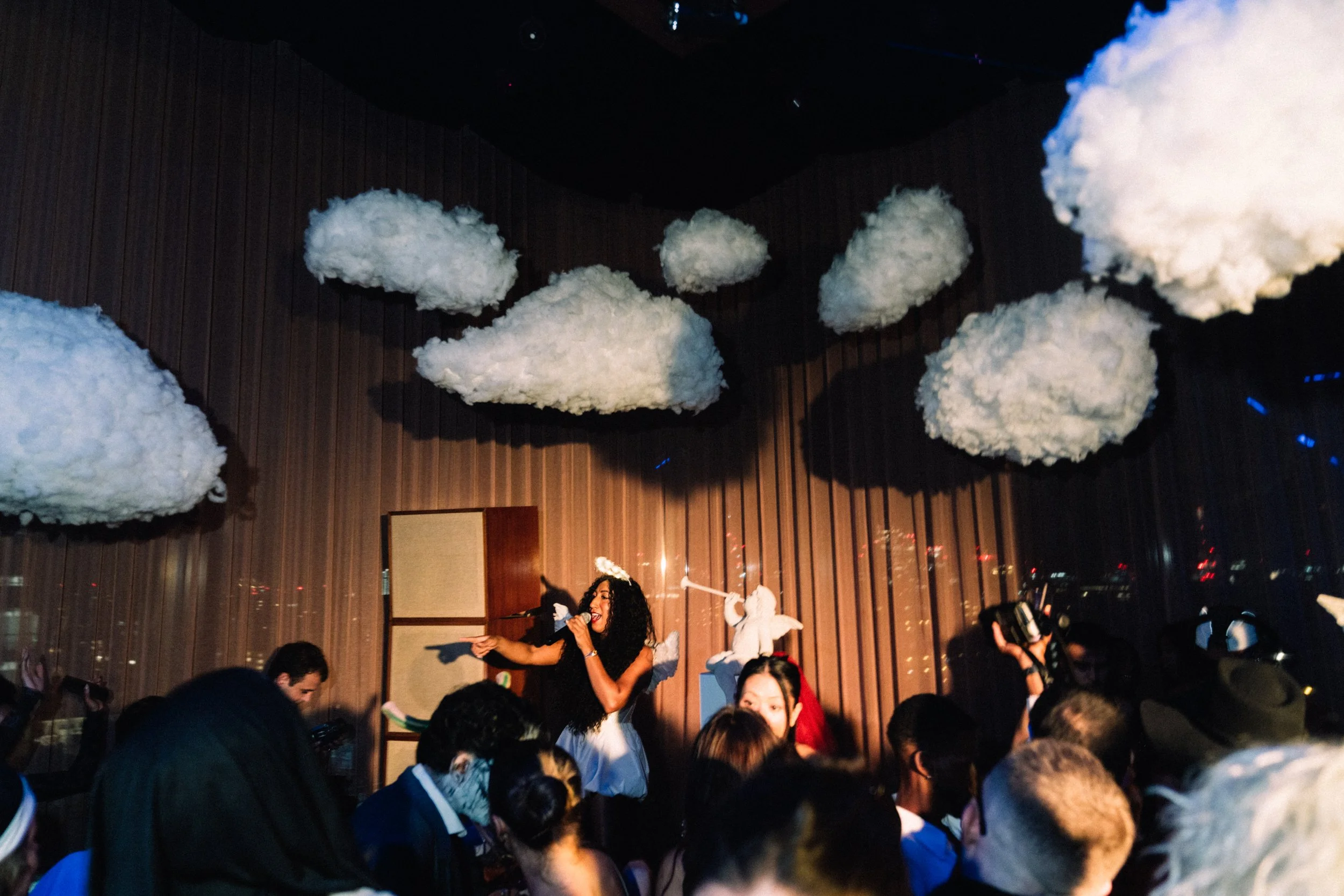 A woman performing on stage surrounded by clouds hanging from the ceiling in a dimly lit venue.