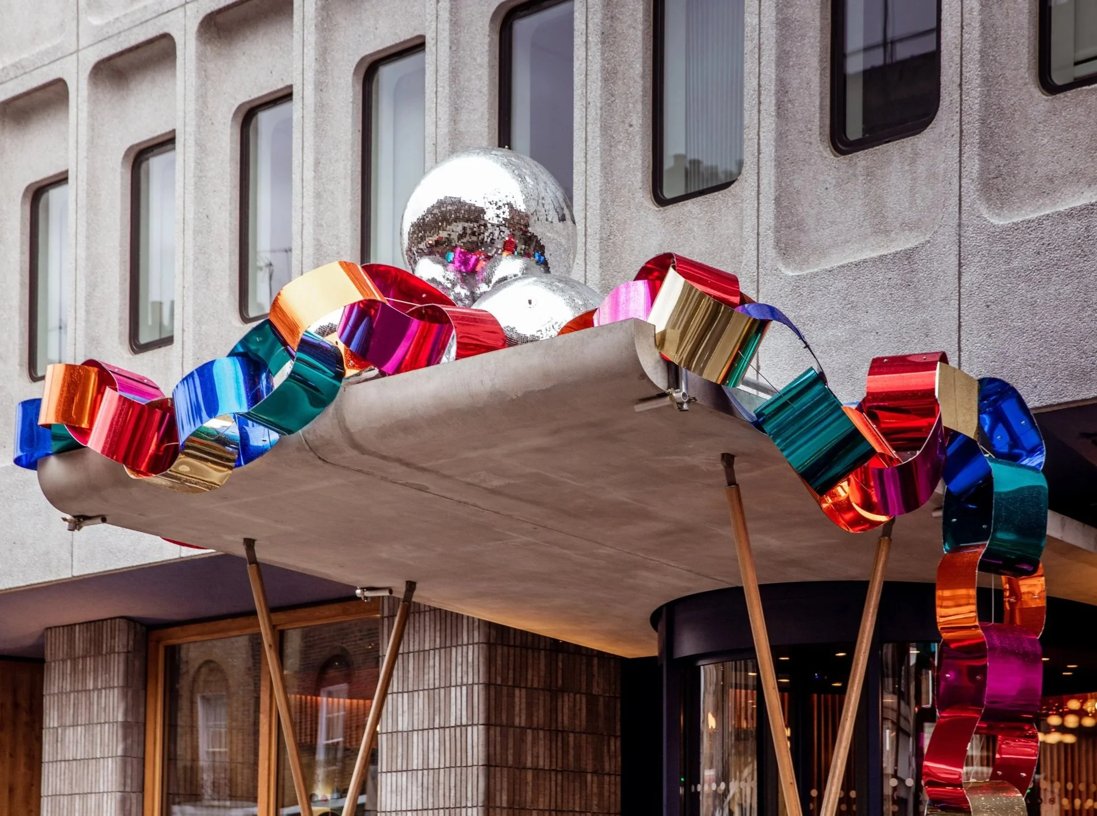 Decorative installation with metallic balloons and colorful ribbons outdoors in front of a building with large windows.