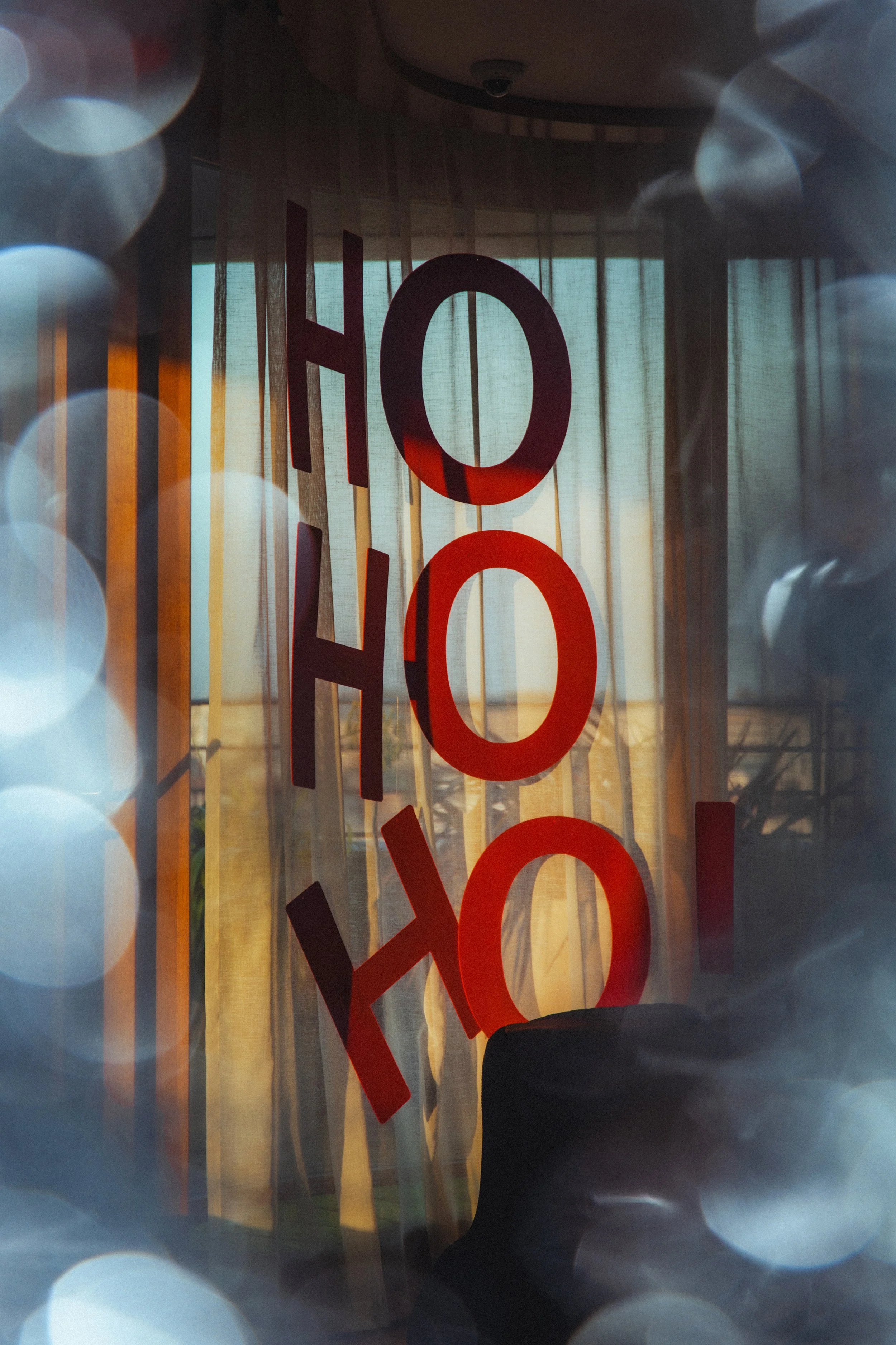 Reflection of a person and large red and black letters on a glass surface with bokeh from lights in the foreground and curtains in the background.