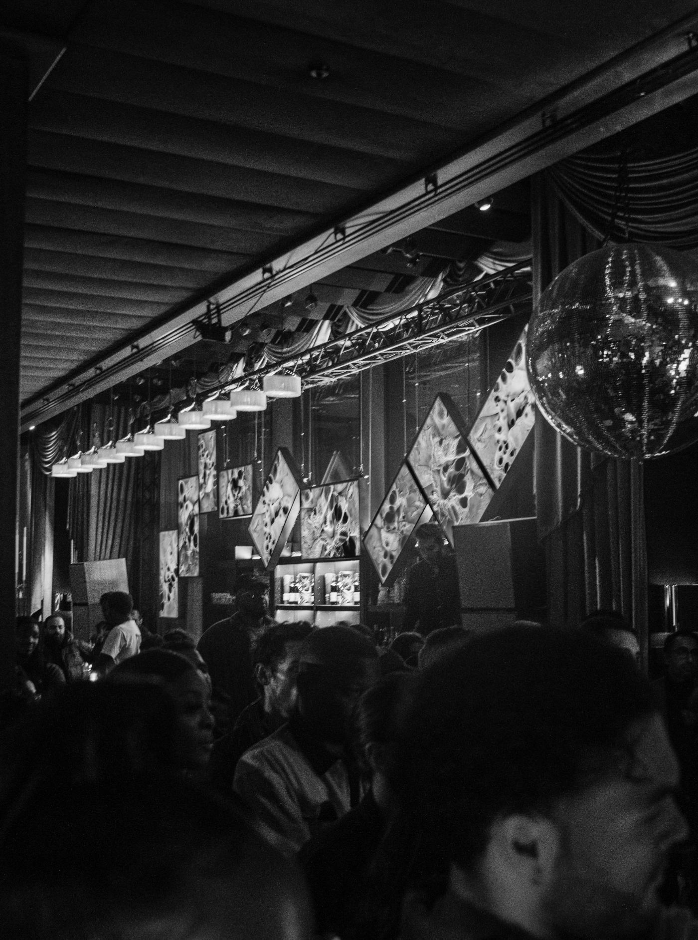 Black and white photo of a crowded indoor event, with many people seated and standing, a large disco ball hanging from the ceiling, and abstract art on the walls.