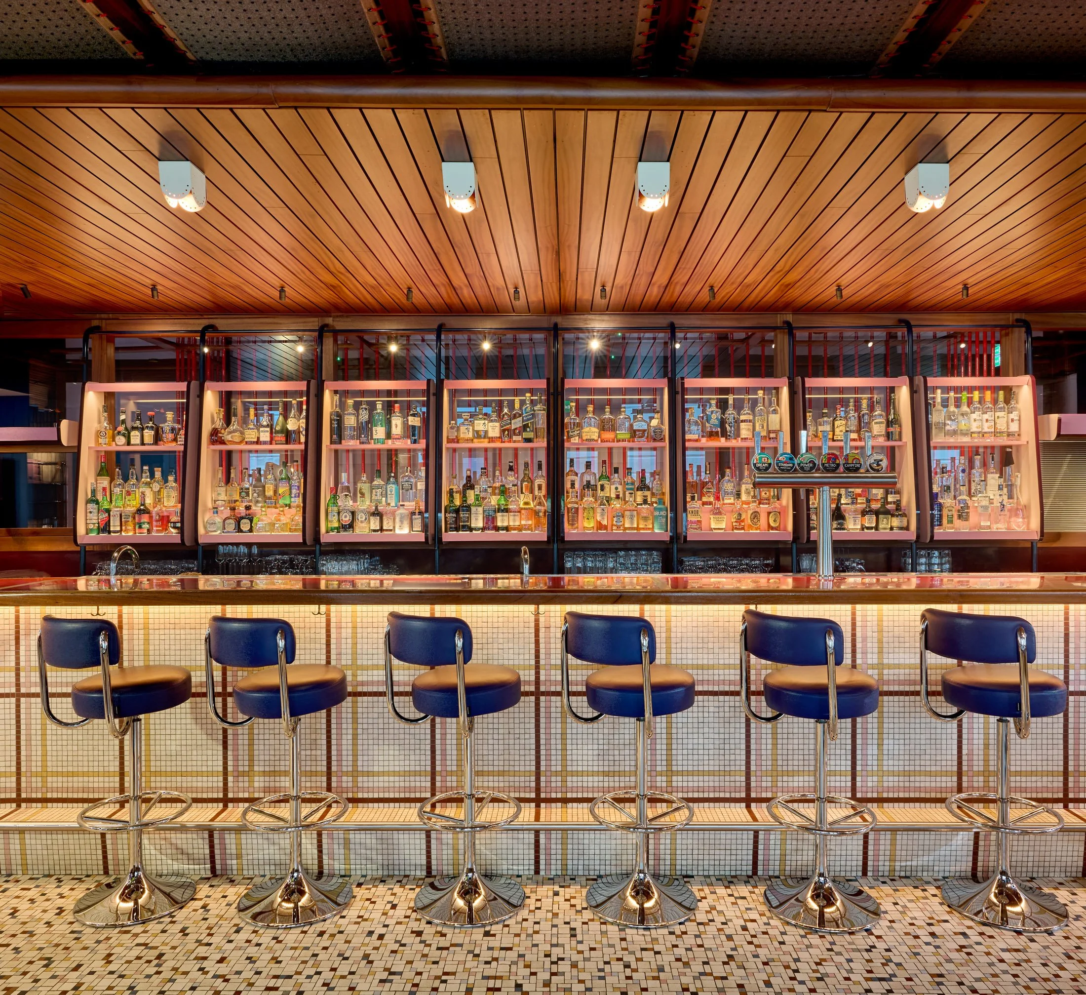 Bar with a wood-paneled ceiling, bottles of alcohol on shelves, bar stools, and a tiled floor.