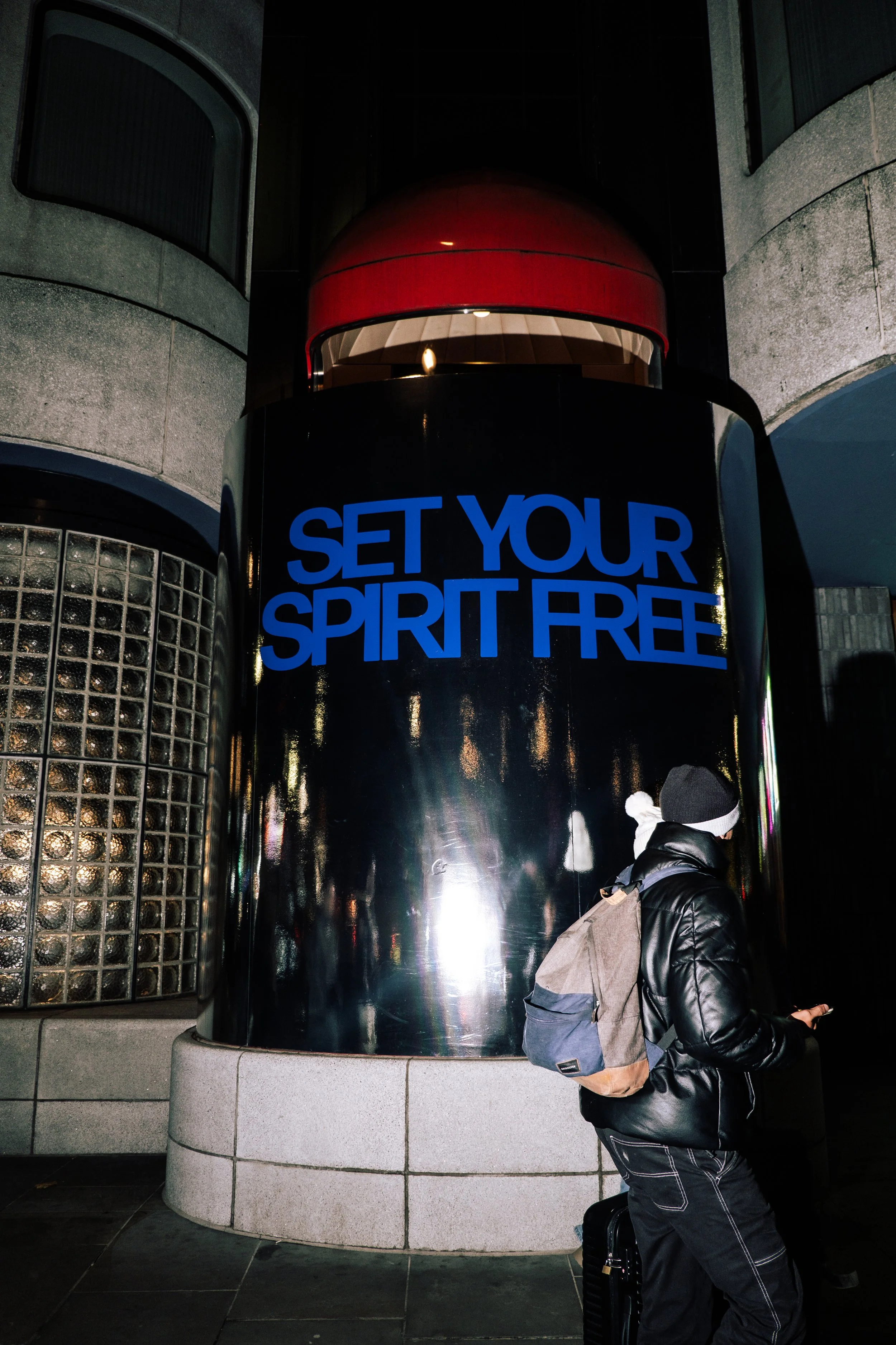 A person with a backpack and coat standing at a modern building entrance, which features a large illuminated sign that reads 'Set Your Spirit Free' and metallic architectural elements.