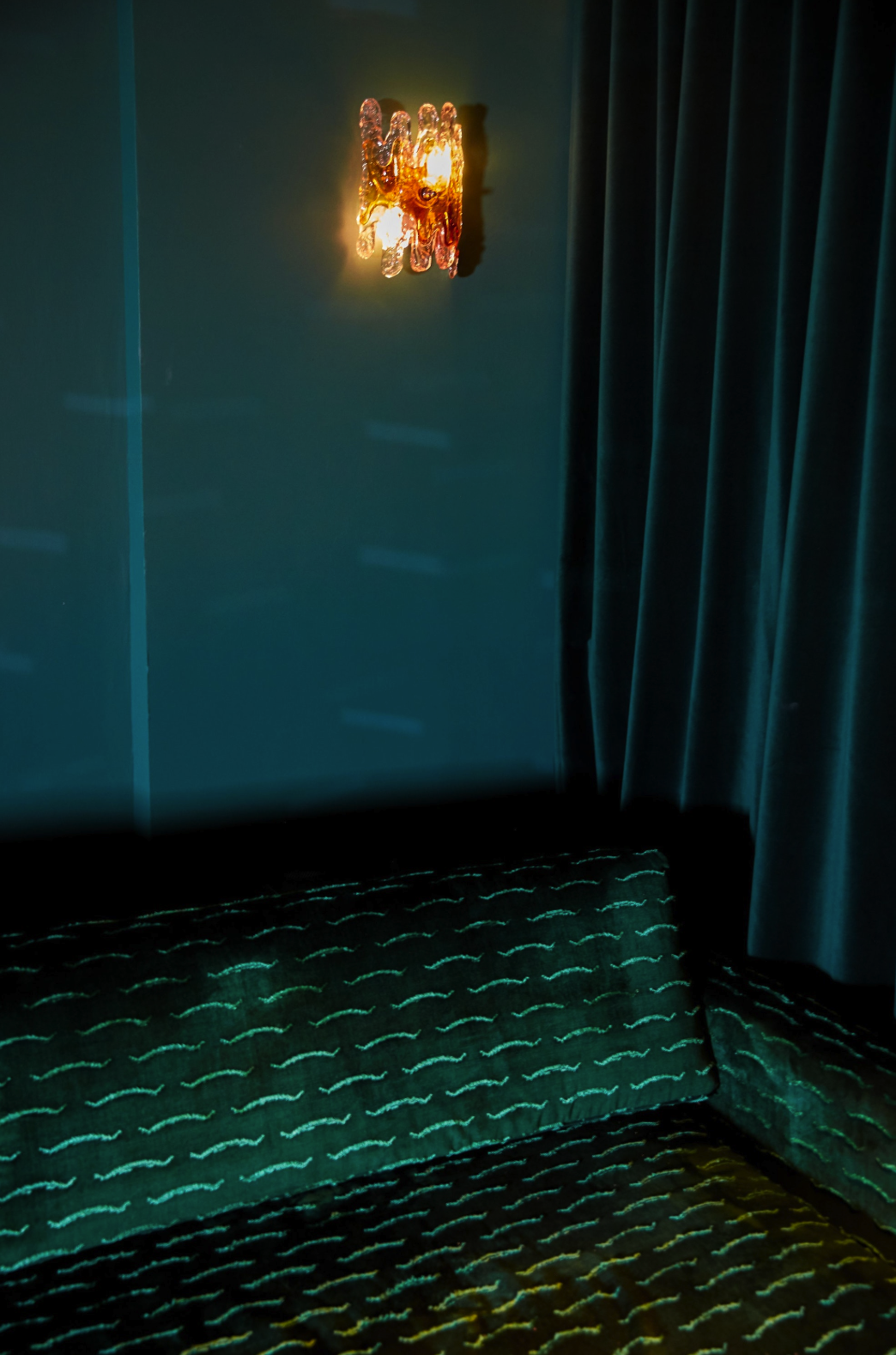 Interior of a room with teal walls, teal velvet curtains, a decorative glowing wall sconce, and a patterned teal and yellow ottoman.