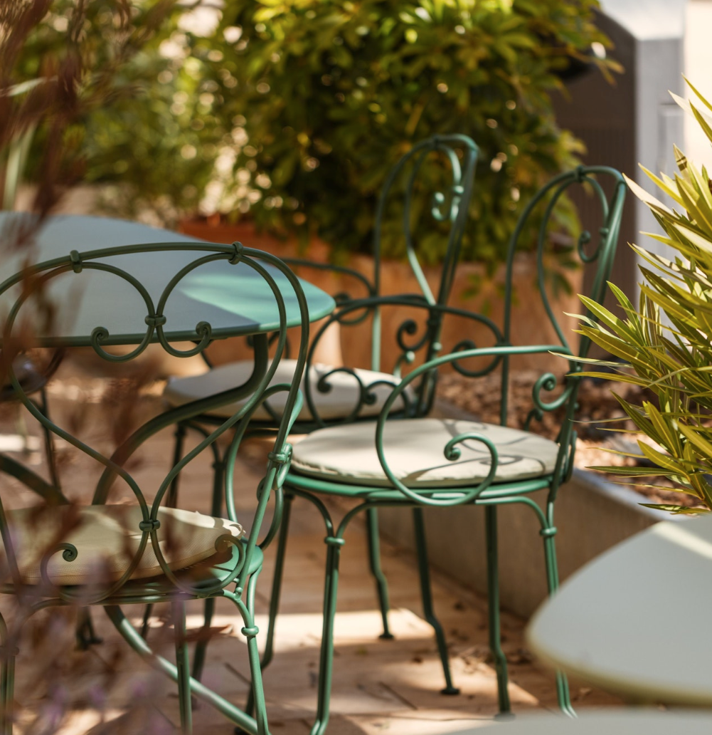 Green metal outdoor table and chairs with white cushions on a patio, surrounded by green plants and bushes.