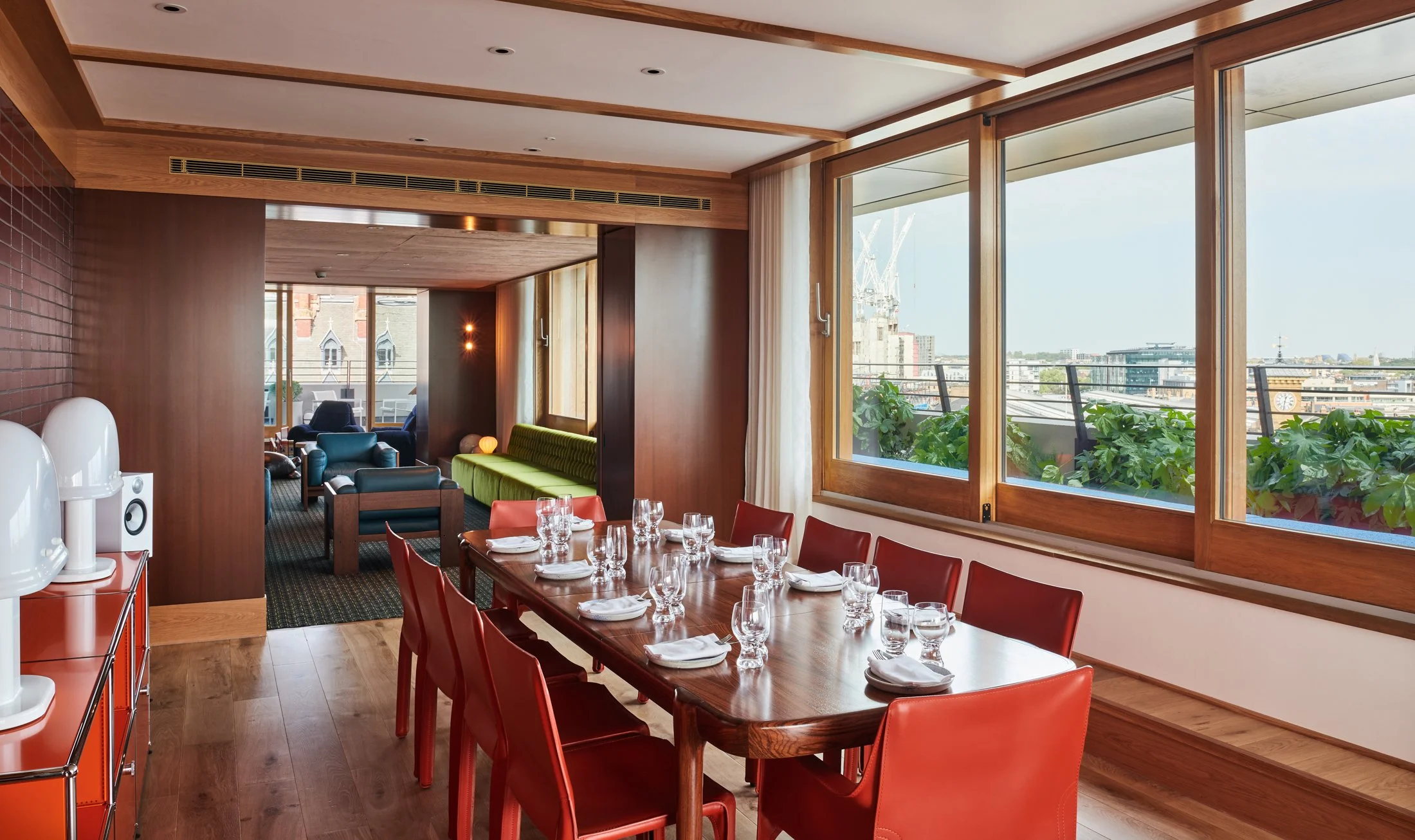 A dining area with a wooden table set with white napkins, glasses, and plates, surrounded by red chairs, with large windows showing a cityscape and a balcony with plants, and a seating area with colorful chairs in the background.