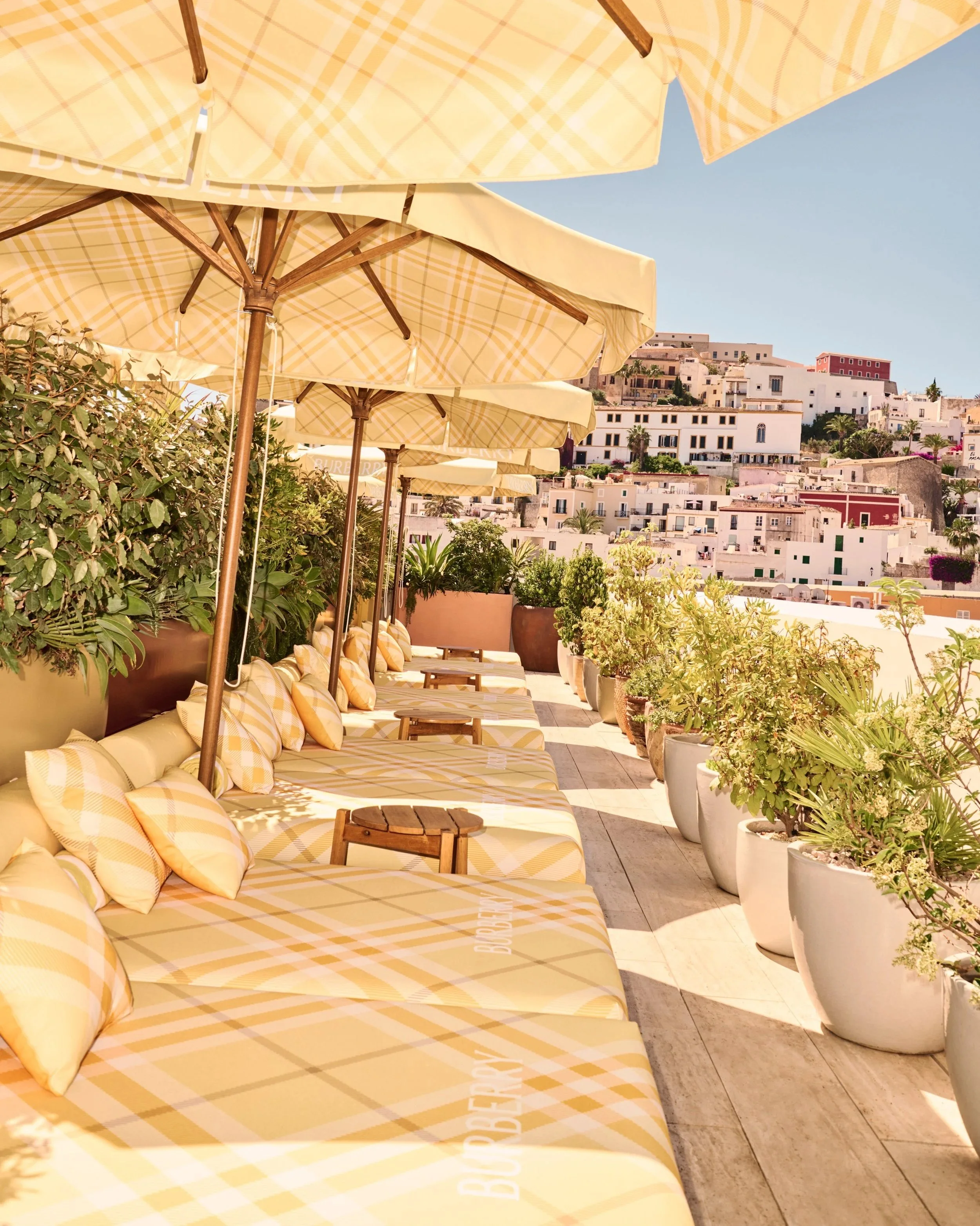 Outdoor seating area with yellow plaid umbrellas, lounge chairs with matching cushions, large white potted plants, and a view of white buildings on a hillside under a clear blue sky.