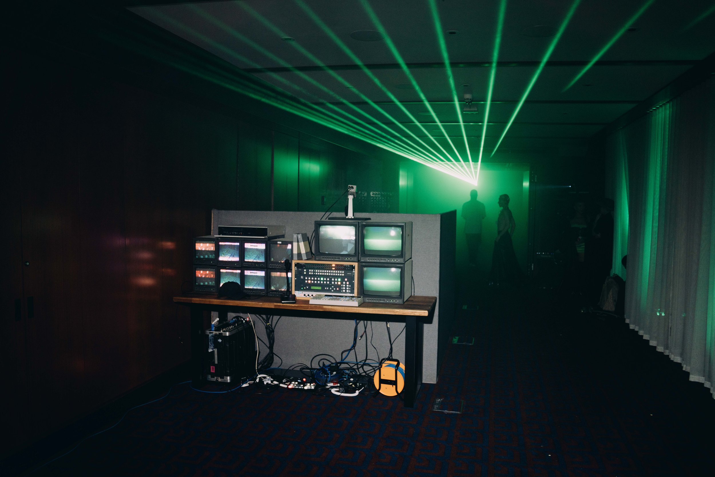 Dark room with green laser lights and several monitors on a desk, with two people standing in silhouette in the background.
