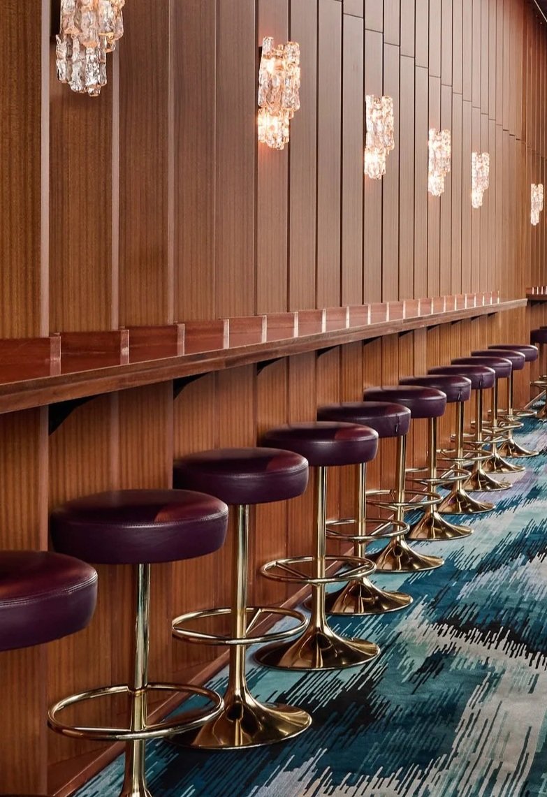 A row of purple bar stools with gold bases lined up against a wooden wall with decorative hanging lights.