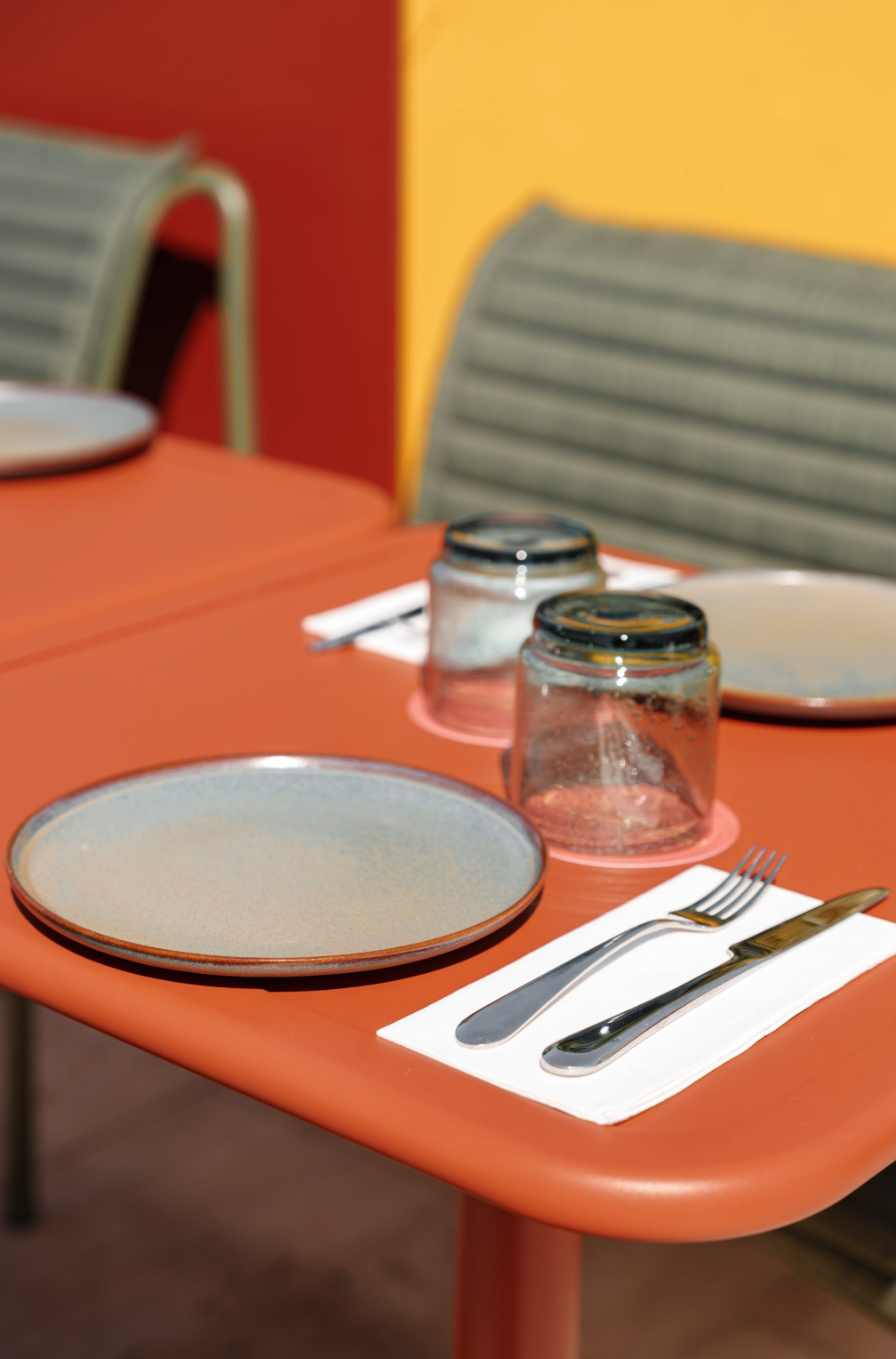 A table set with two ceramic plates, two upside-down glass cups, a fork and knife on a white napkin on an orange table with colorful walls and chairs in the background.