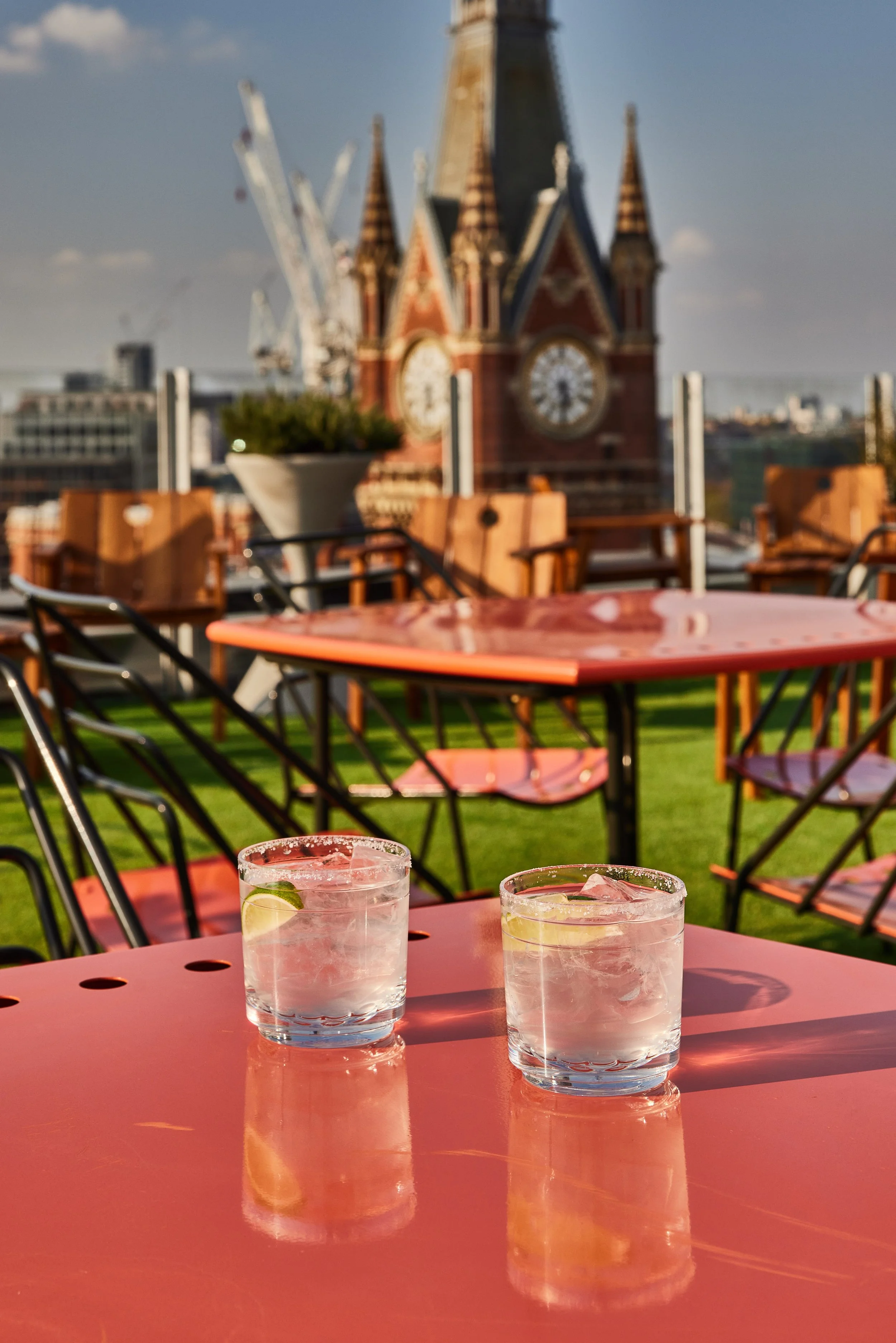 Two glasses of water with lemon slices and ice on a pink table, outdoor setting with chairs and a large clock tower in the background.