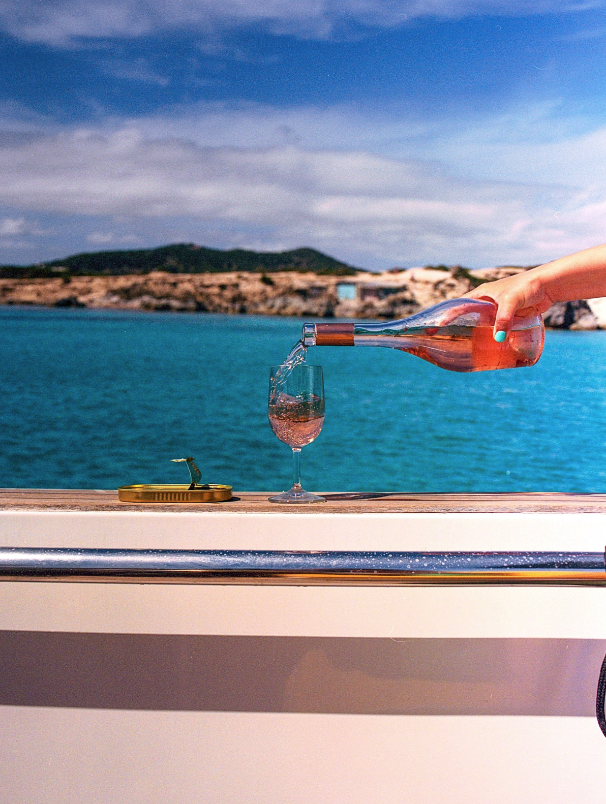 Person pouring rosé wine from a bottle into a glass on a boat deck by the water with a coastal landscape and blue sky in the background.