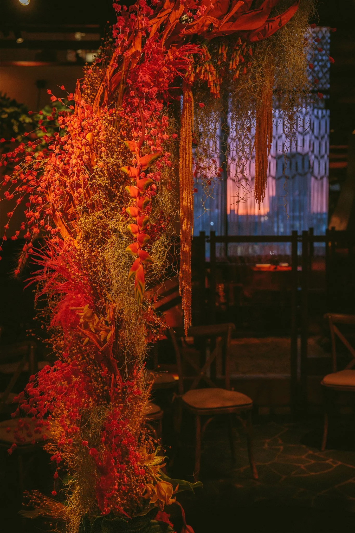 A floral arrangement featuring red, orange, and yellow flowers and greenery, set in a dark interior with wooden furniture and a geometric patterned window in the background.