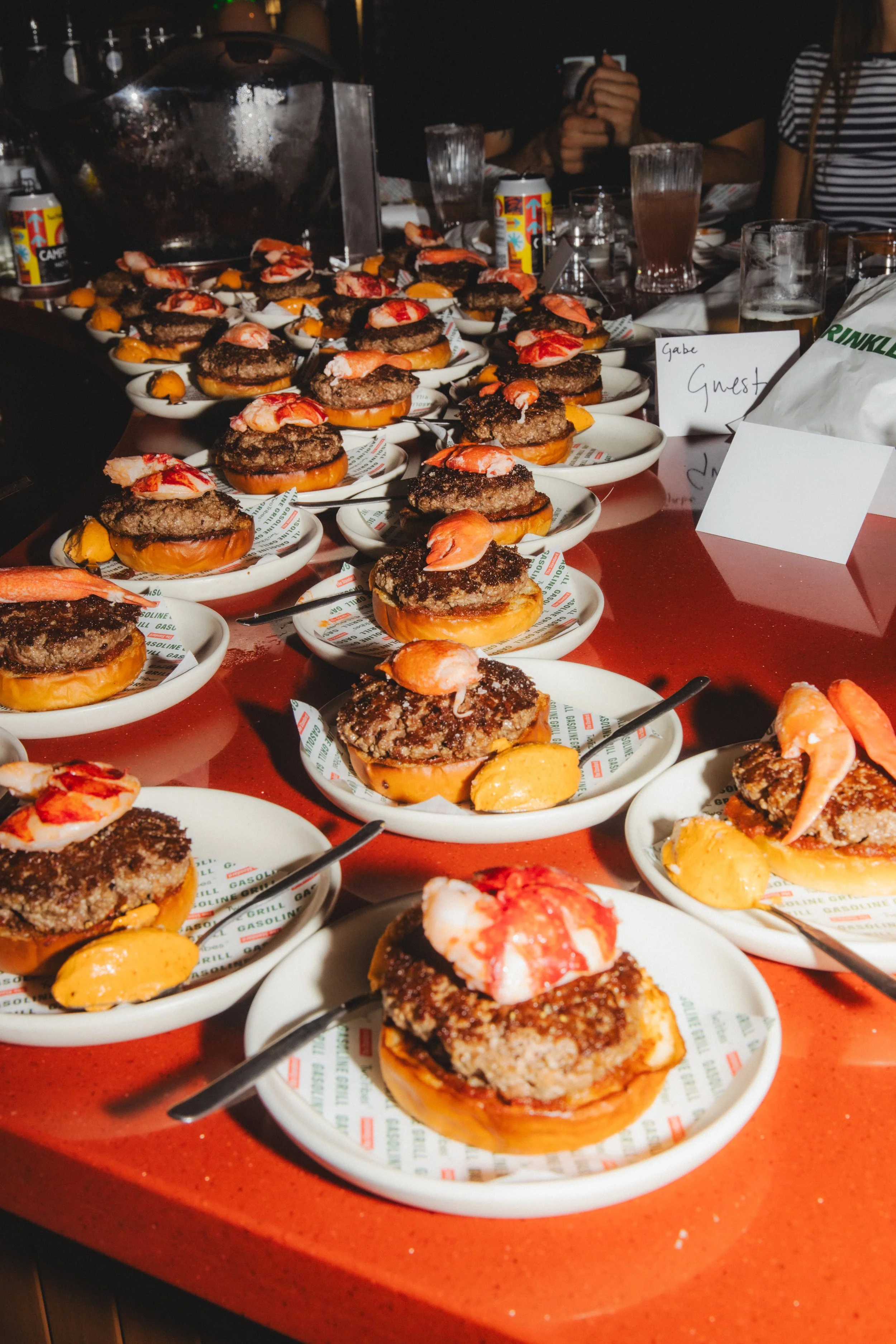 Multiple mini burgers with shrimp, sauce, and toppings on white plates arranged on a red table, with people in the background.