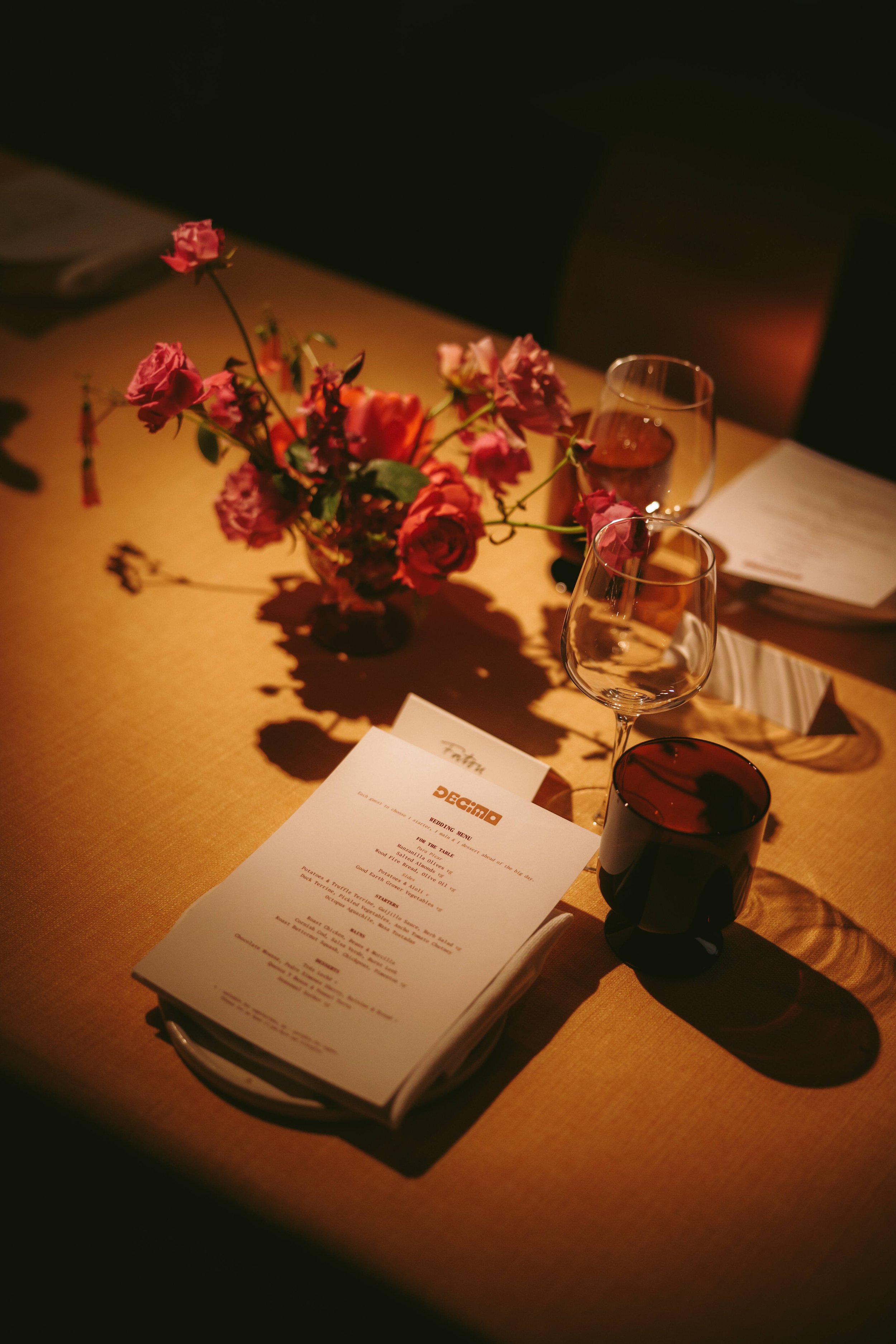 A dimly lit table with a vase of pink and red flowers, two glasses of wine, and a menu booklet.