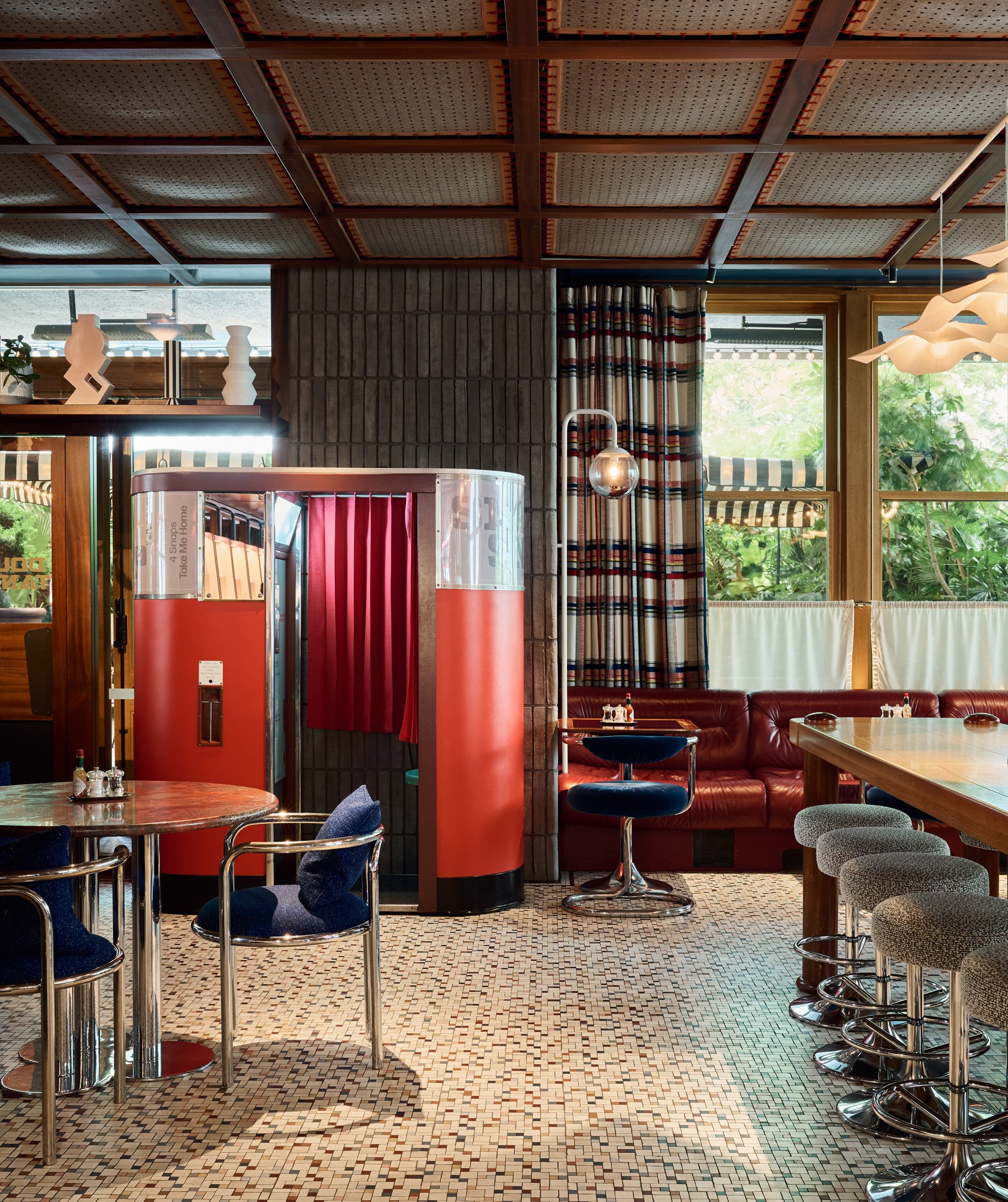 Interior of a restaurant or cafe with patterned tile flooring, wooden ceiling beams, large windows with striped curtains, and various chairs and tables, including a red booth and bar stools.
