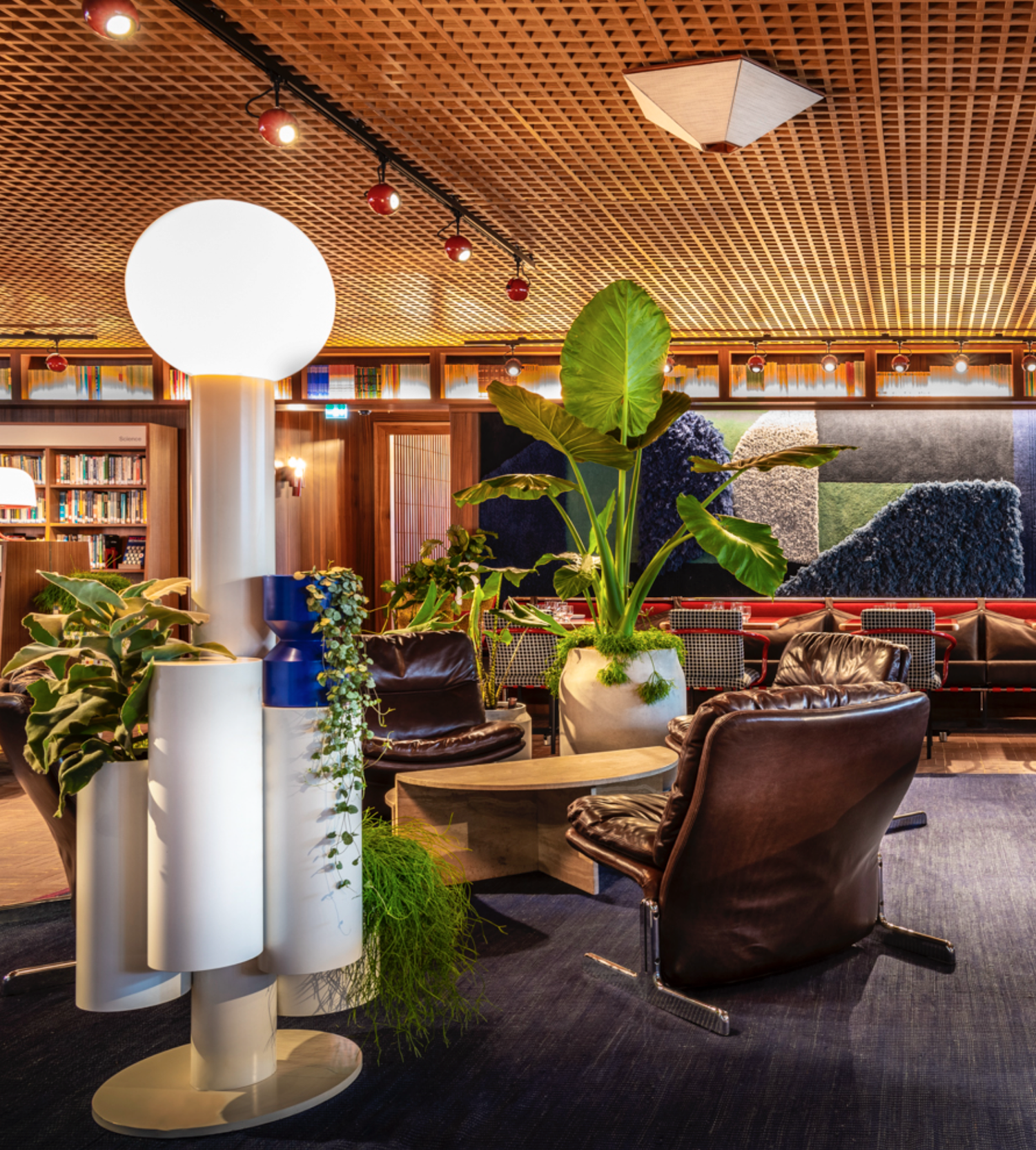 Interior of a modern lounge or library area with leather chairs, potted plants, wooden bookshelves, and colorful wall art, illuminated by warm lighting and ceiling lights.