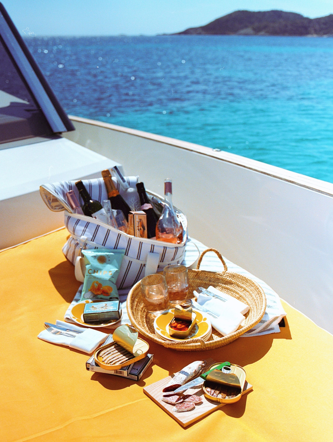 A sunny outdoor table set on a boat with drinks, snacks, and utensils, overlooking blue ocean water and a distant island.