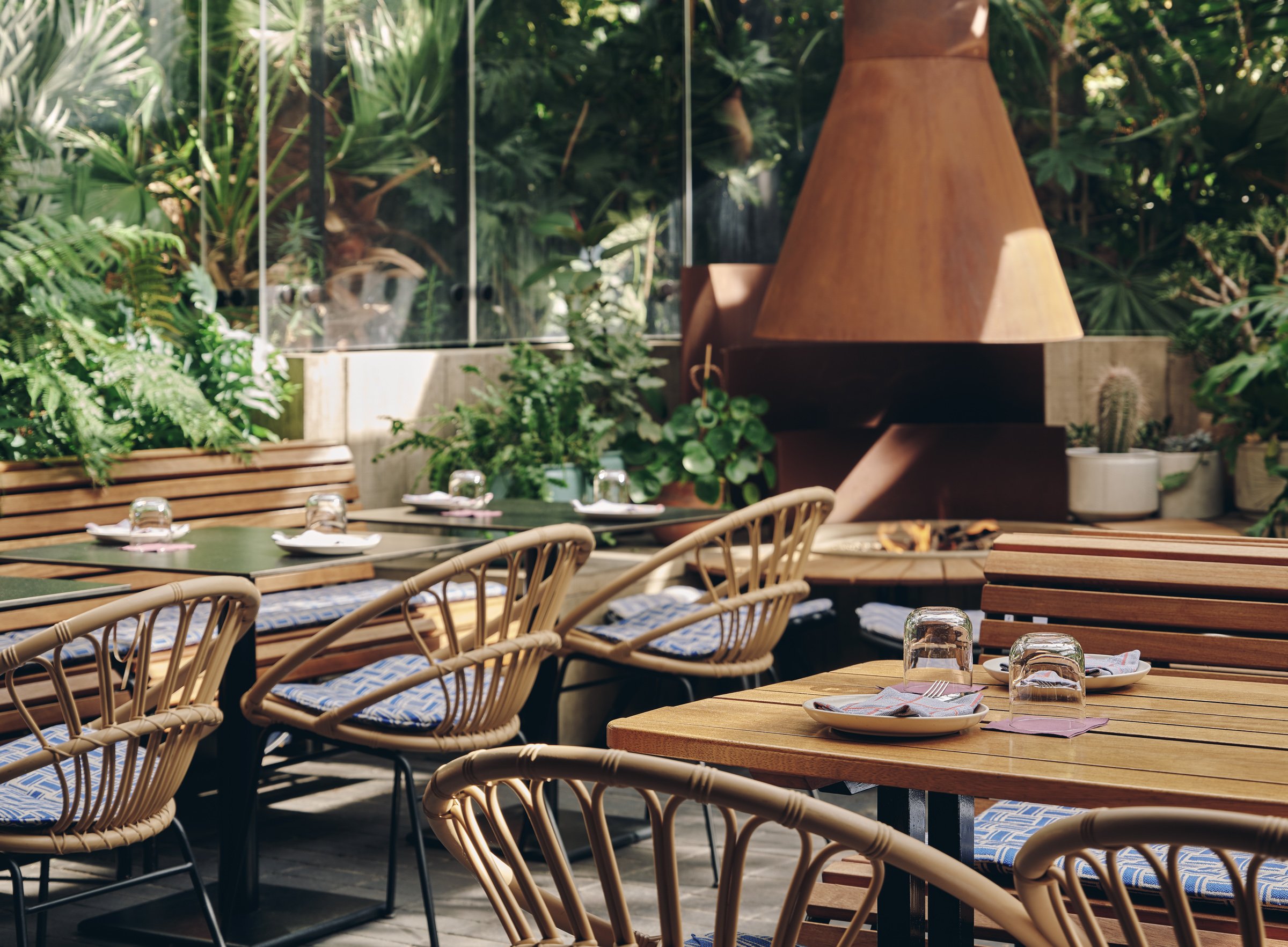 Indoor restaurant with wooden tables set with plates, forks, napkins, and glasses, surrounded by wicker chairs with blue and white cushions, large windows, and lush green plants outside.