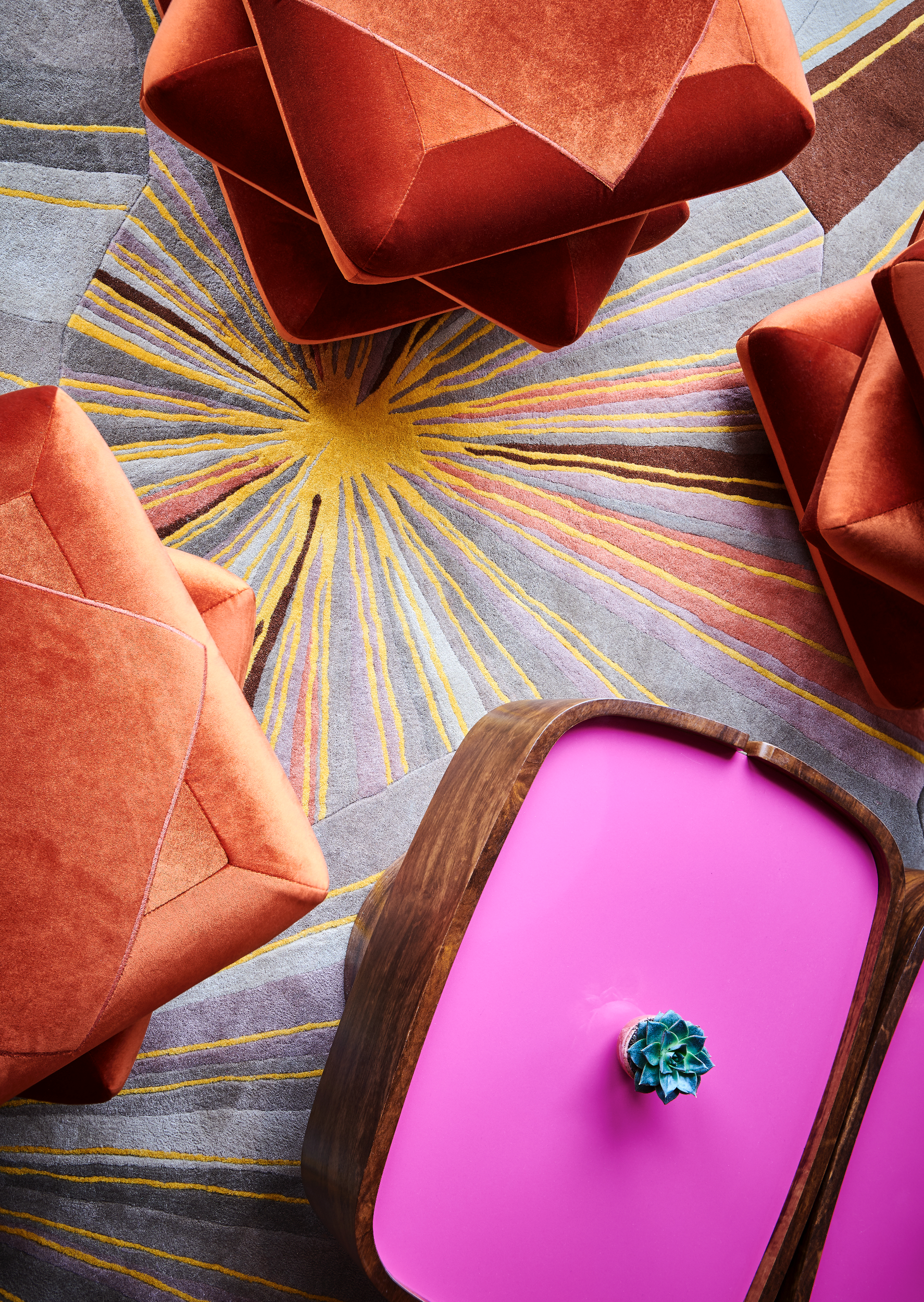 Top down view of a pink cushioned chair with a succulent plant on it, next to two orange velvet armchairs, and a colorful patterned rug.