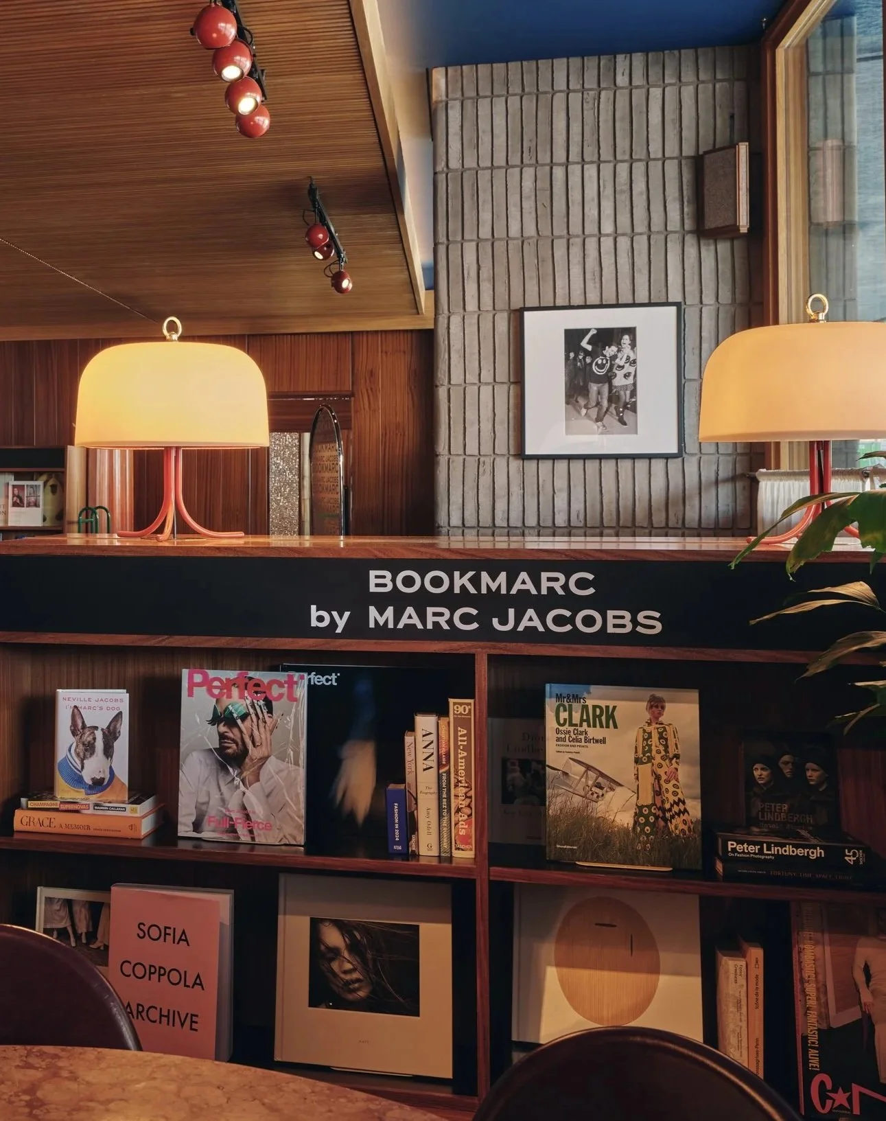 Interior of a bookstore or library section with books on display, illuminated by warm yellow lamps, and a framed black-and-white photograph on a brick wall. A sign reads 'BOOKMARc by MARC JACOBS'.