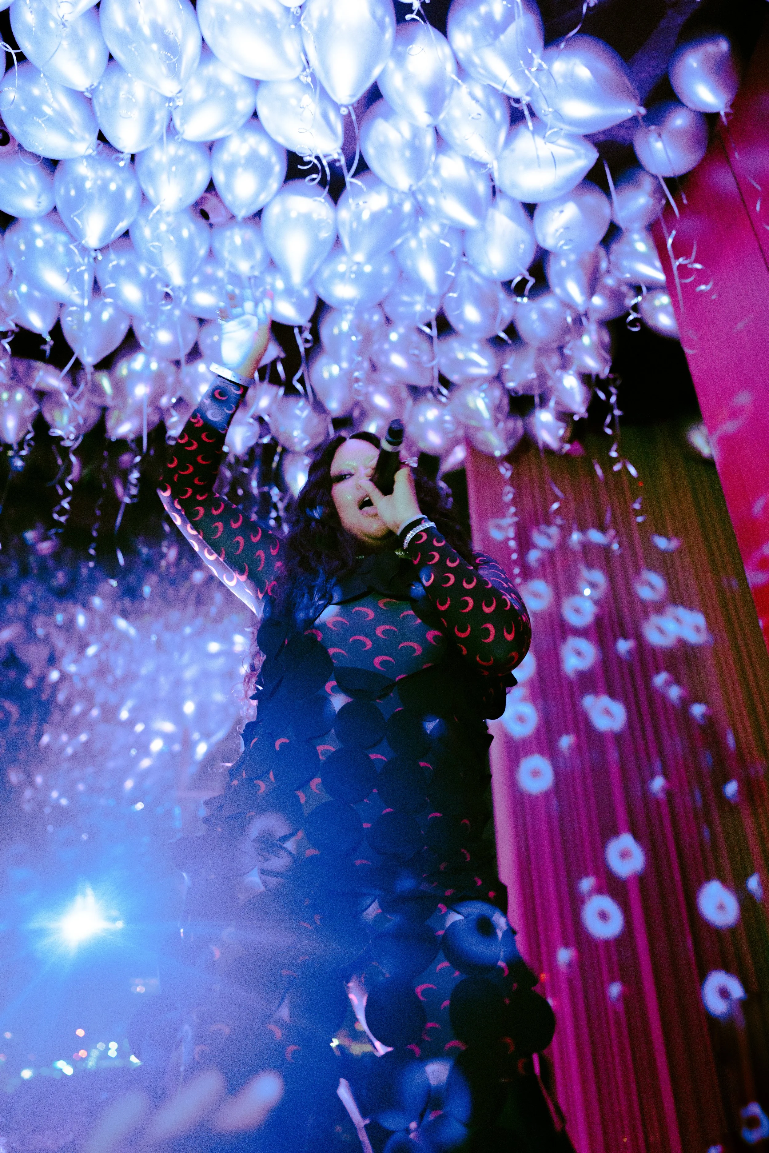 A woman with dark hair and a black outfit with pink moon patterns is celebrating among white balloons with curly ribbons hanging from them, illuminated by pink and purple lighting in a festive setting.
