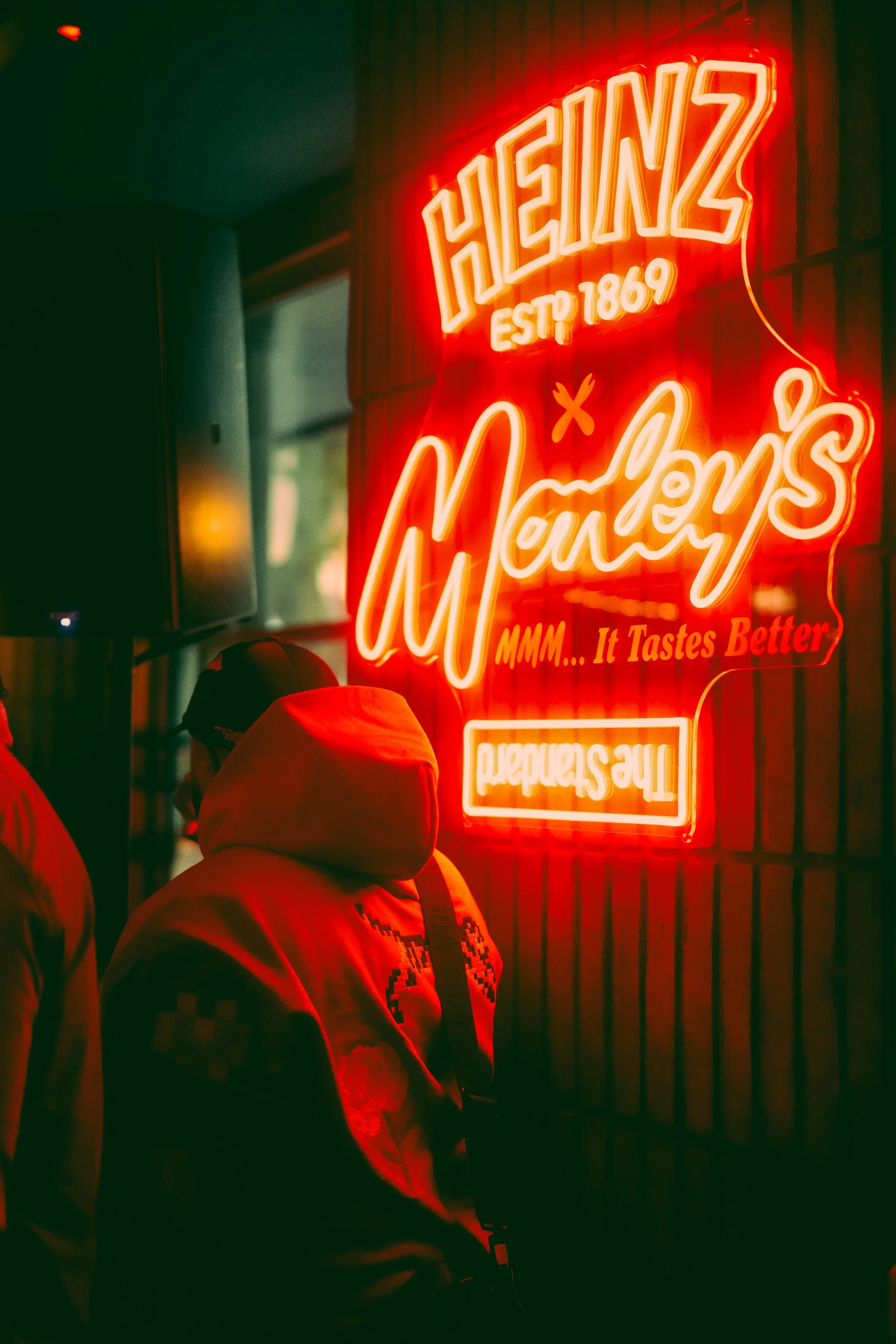 People standing in front of a neon sign that reads 'Heinz EST 1869' and 'MMM... It Tastes Better' at night.