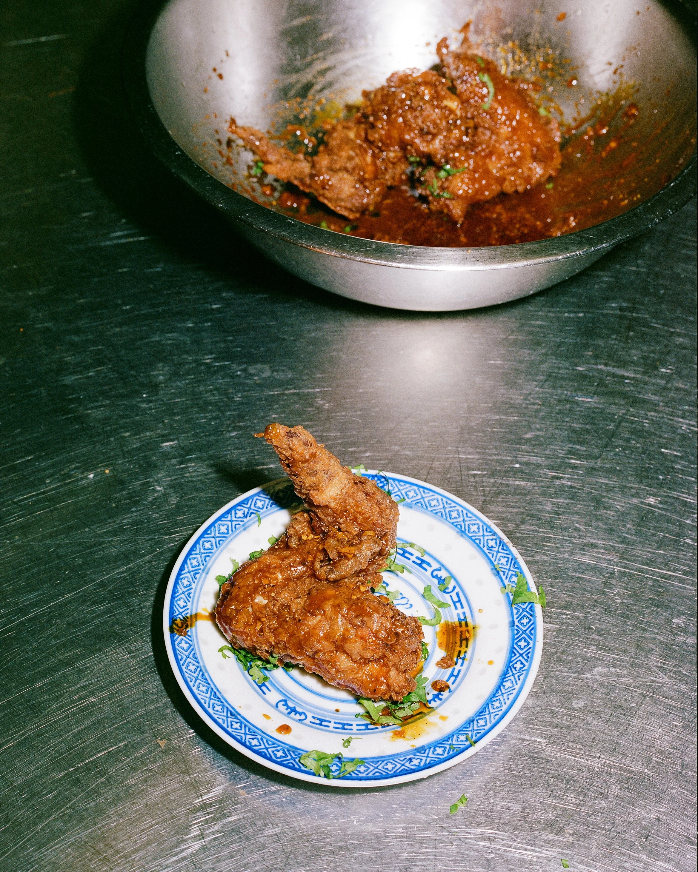 Close-up of a plate with a fried fish fillet covered in brown sauce, placed on a decorative blue and white plate on a metal table. In the background, there is a metal bowl with a serving of spicy meat in sauce.