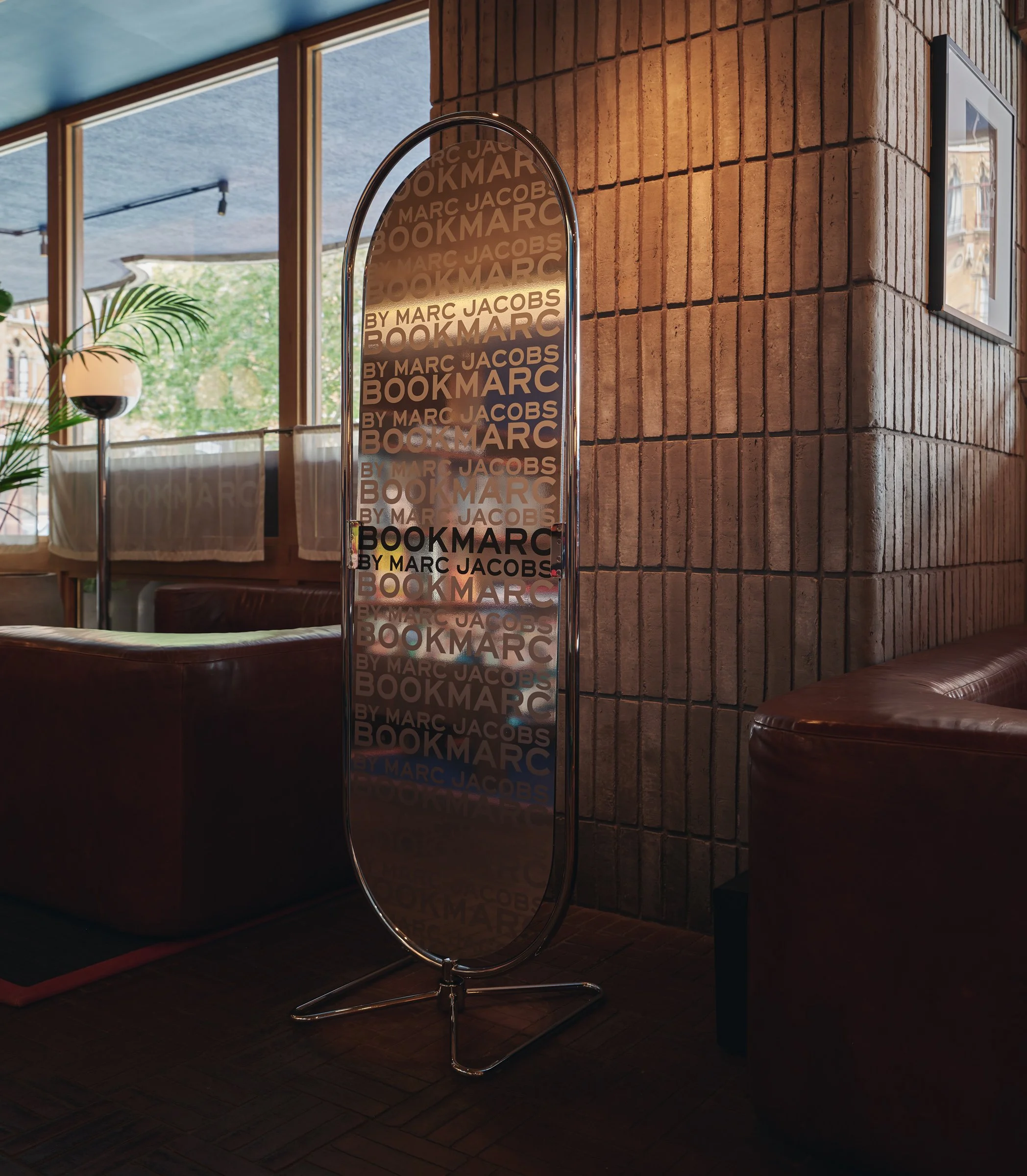A tall, oval-shaped mirror with a metal frame and stand, displaying the repeated text 'BY MARC JACOBS BOOKMAR' in gold and black, in a room with a brick wall, wooden floor, large window with curtains, and some indoor plants.