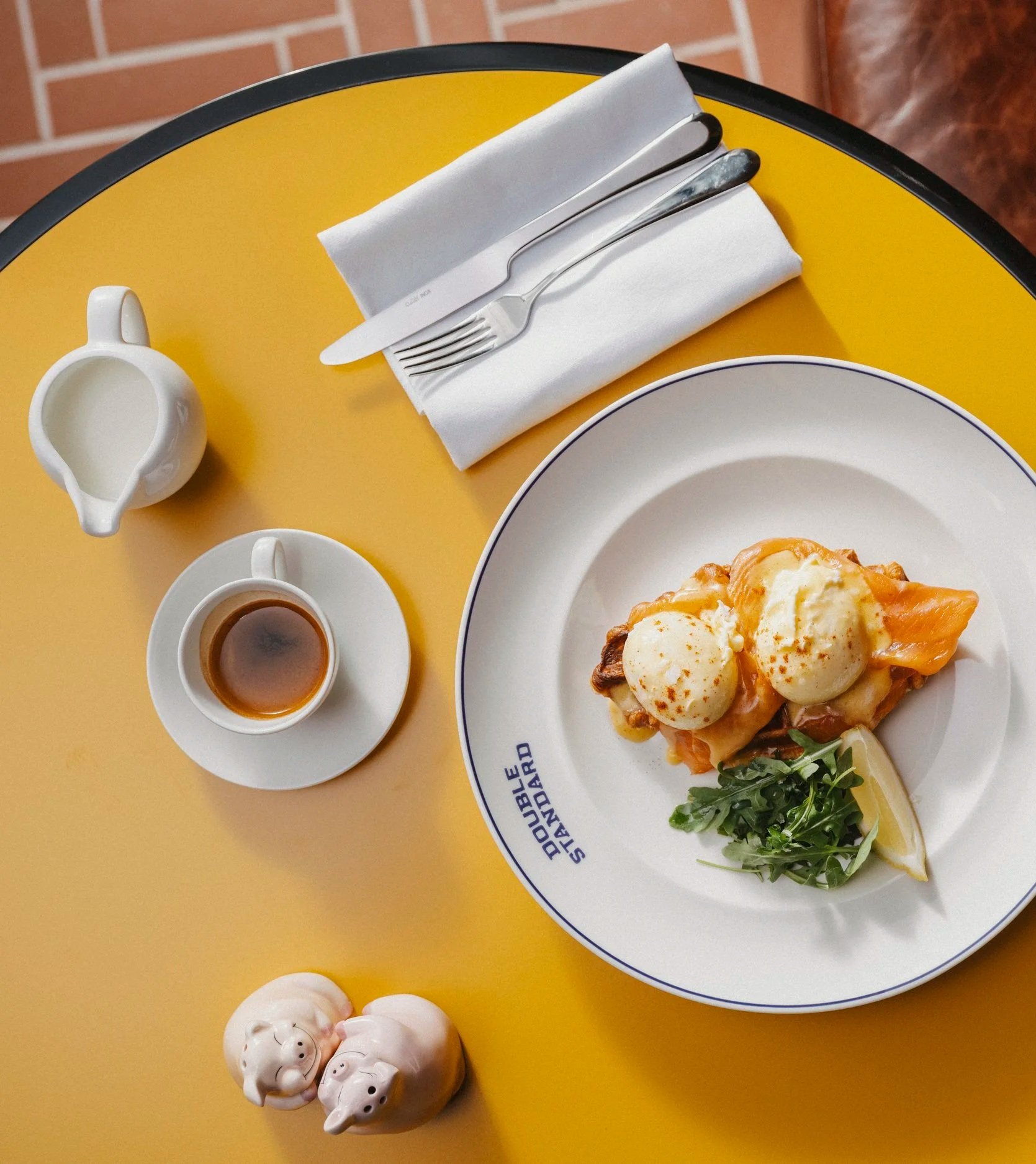 A breakfast plate featuring eggs Benedict, a small pitcher of syrup, a cup of syrup, and four pig-shaped sugar containers on a yellow table, with utensils and napkins.