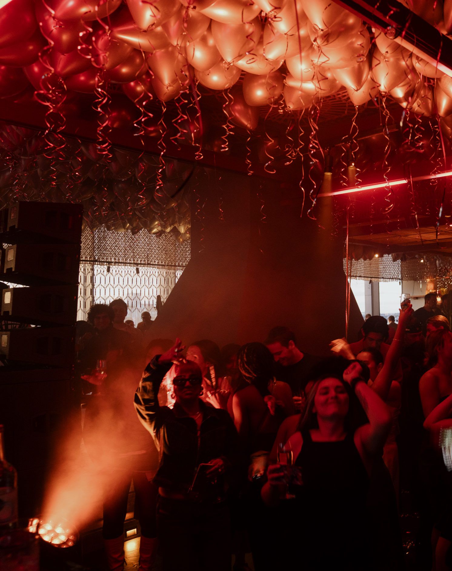 Crowd of people celebrating at a party with red and gold balloons hanging from the ceiling, people holding drinks and dancing in a dimly lit room with red lighting.