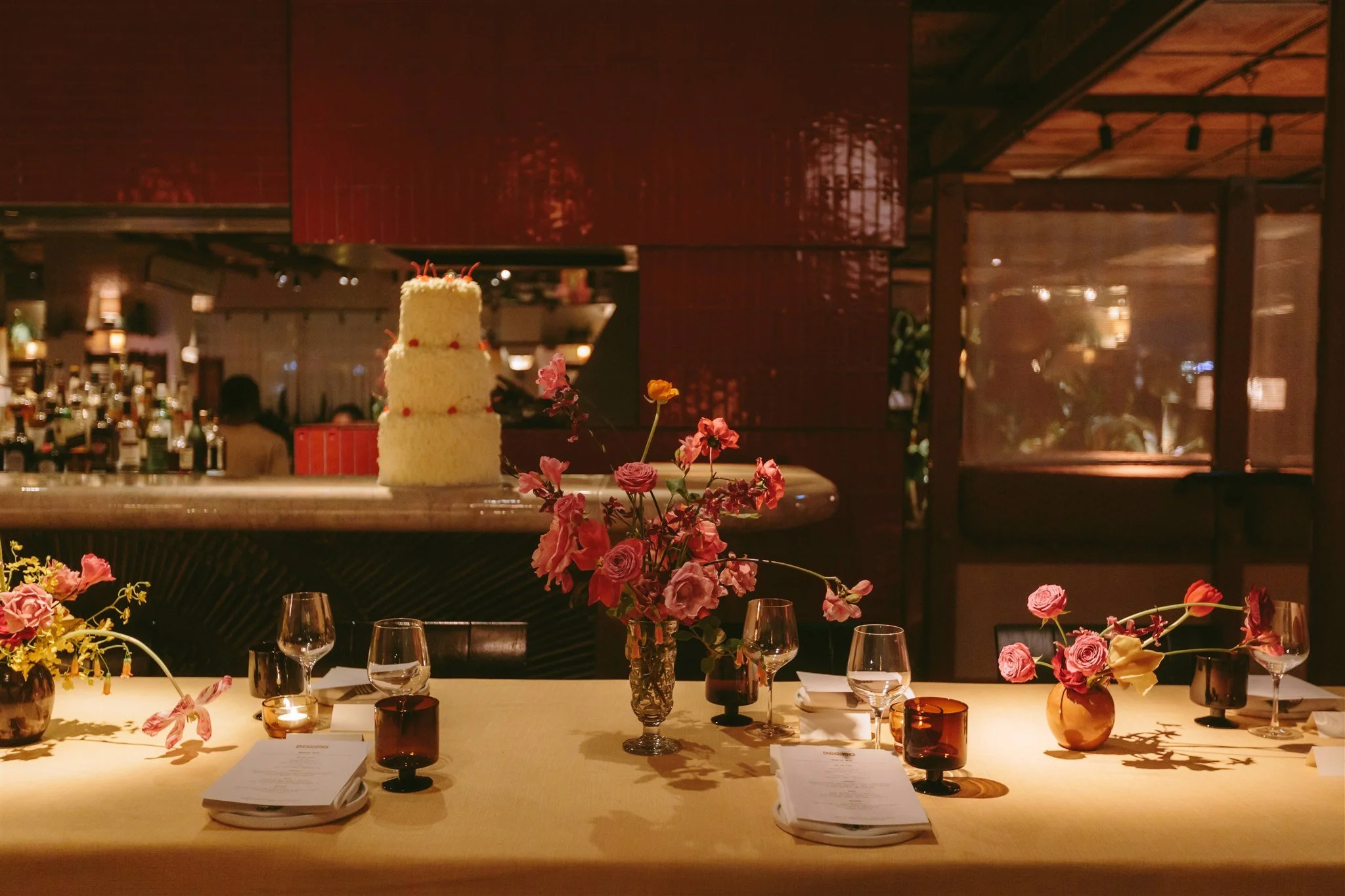 A decorated dining table with vases of pink and yellow flowers, multiple candles, and menus, set in a dimly lit restaurant with a large three-tiered cake on a display in the background and a bar area.