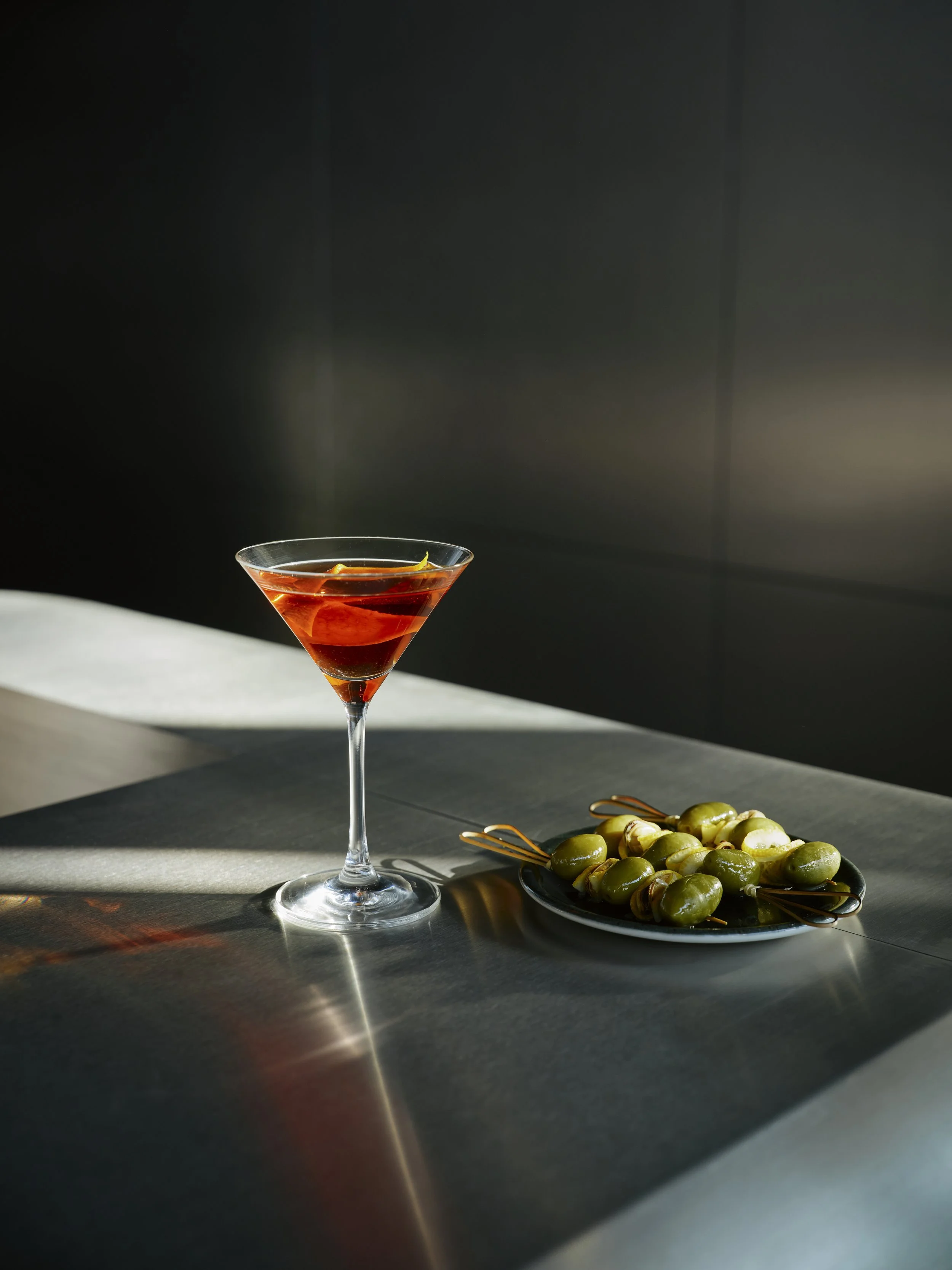 A martini glass filled with an amber-colored cocktail, garnished with a lemon peel, on a metallic surface with a dark background. A plate of green olives with toothpicks is beside the glass.