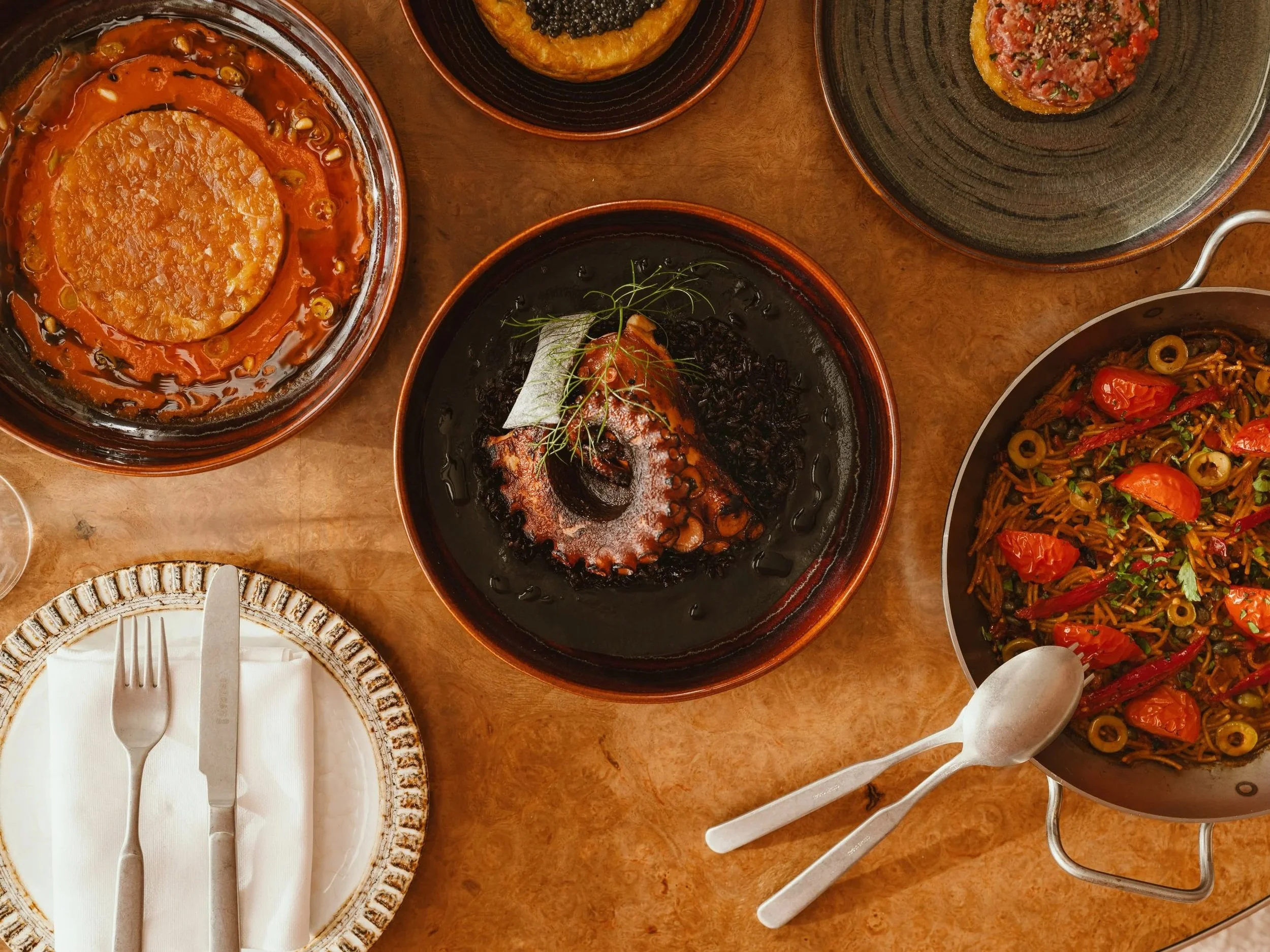A table with various dishes including a large bowl of stew with a piece of bread, a blackfish dish garnished with herbs, a bowl of pasta with cherry tomatoes and olives, and a plate with bread, fork, and knife.