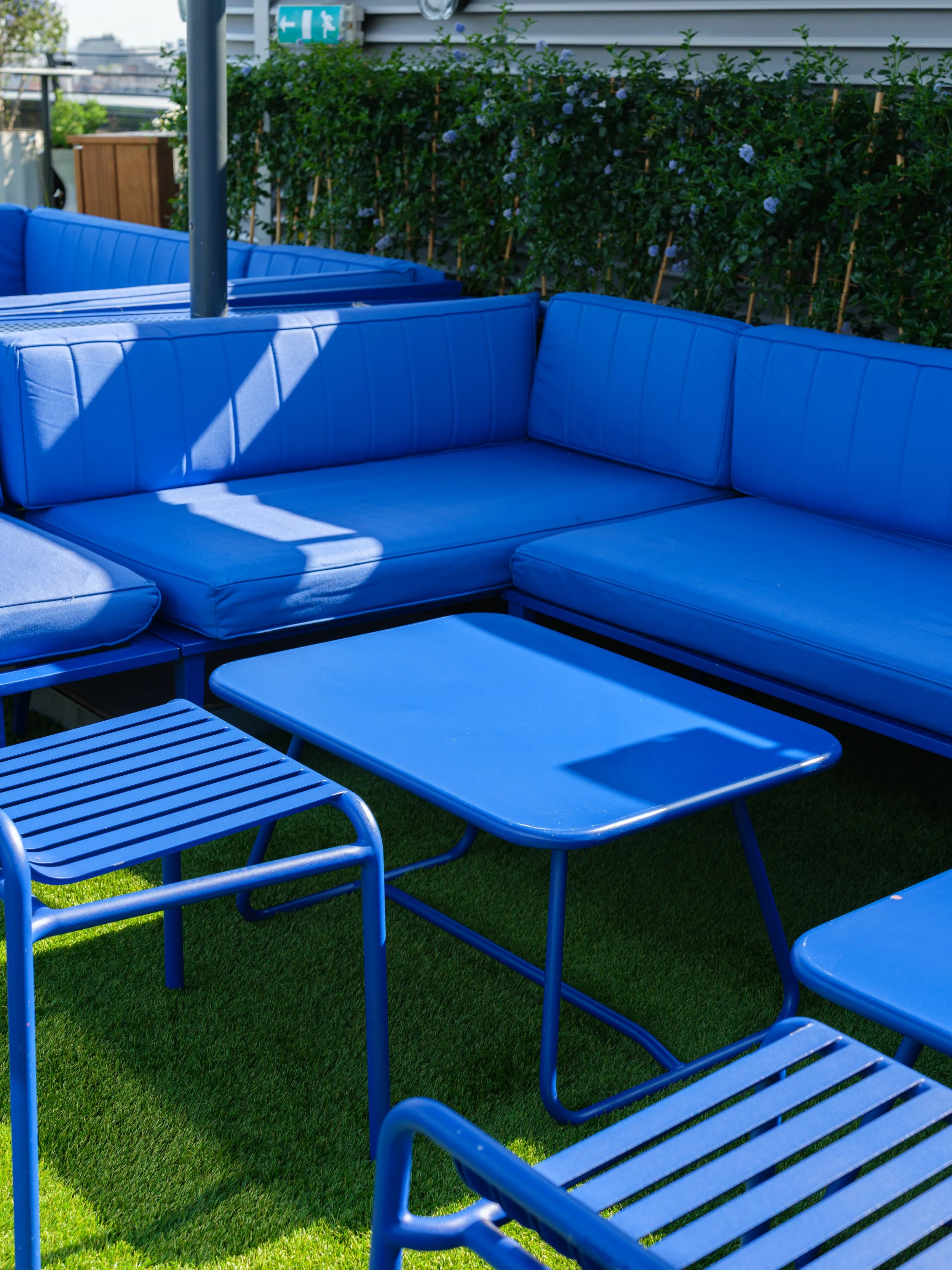 Blue outdoor sectional sofa and matching blue coffee table on a grassy rooftop with green shrubbery and a house wall in the background.