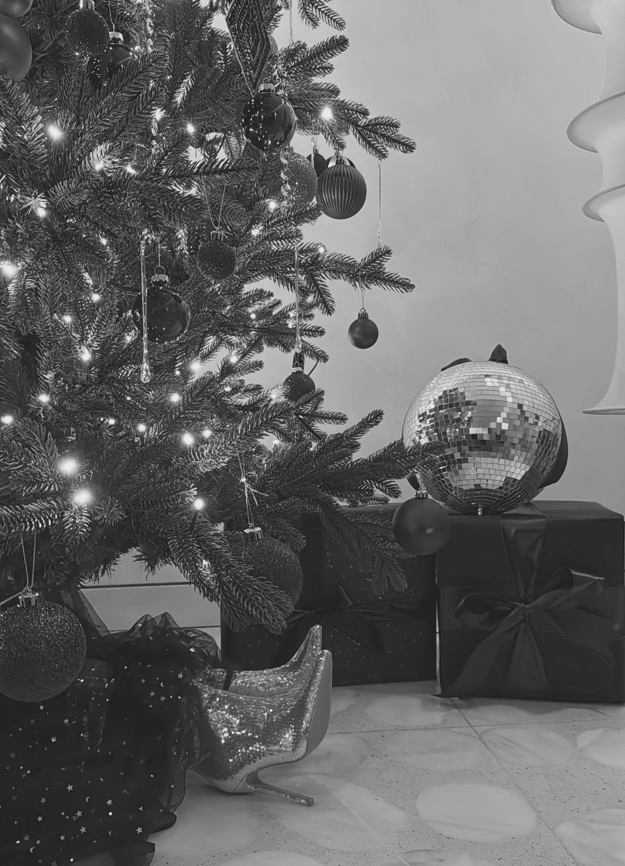 A decorated Christmas tree with lights and ornaments, sitting next to a wrapped gift with a shiny surface and ribbons, in a room with tiled floor and wrapped presents nearby.