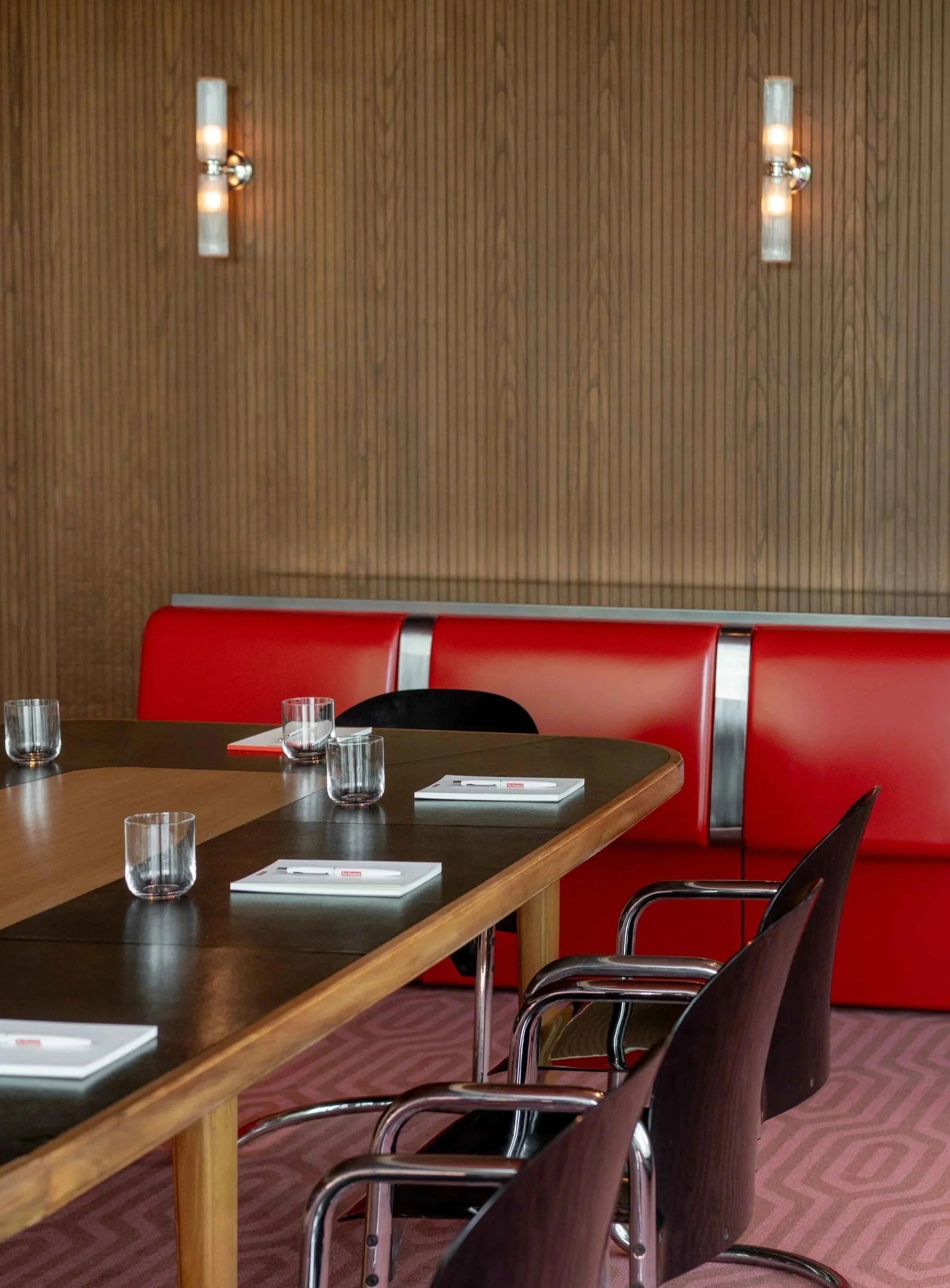 A conference room with a wooden wall, two modern wall sconces, a large oval wooden table set with glasses and notepads, and red cushioned chairs with metal armrests.