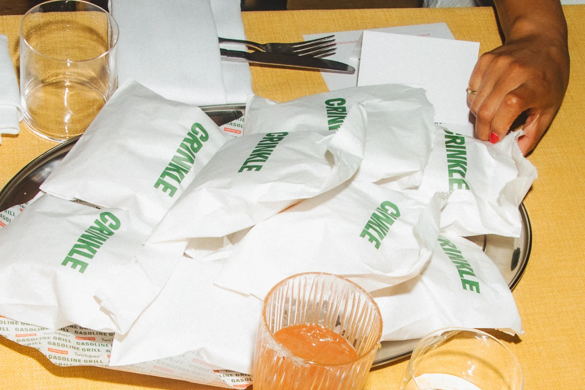A table with crumpled white paper liners that have green writing, a glass of iced tea with lemon, a fork and knife on a white napkin, and a person’s hand with red nail polish resting on the table.