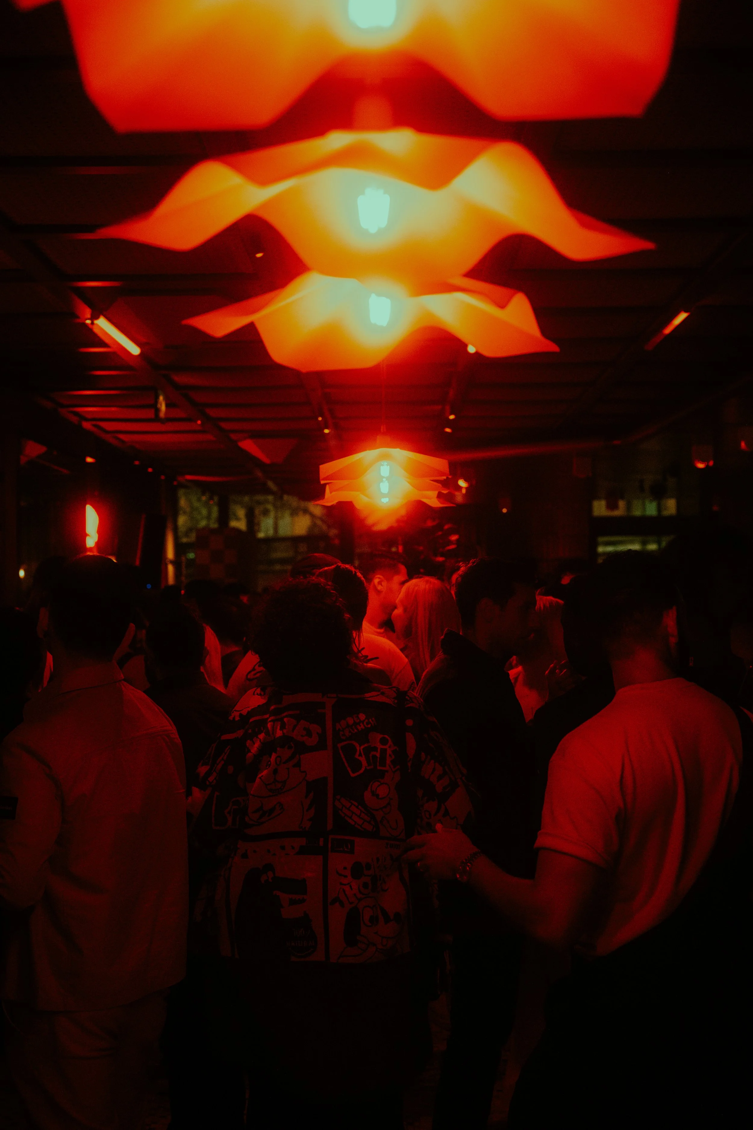 A group of people standing under orange and yellow geometric hanging light fixtures in a dimly lit room.