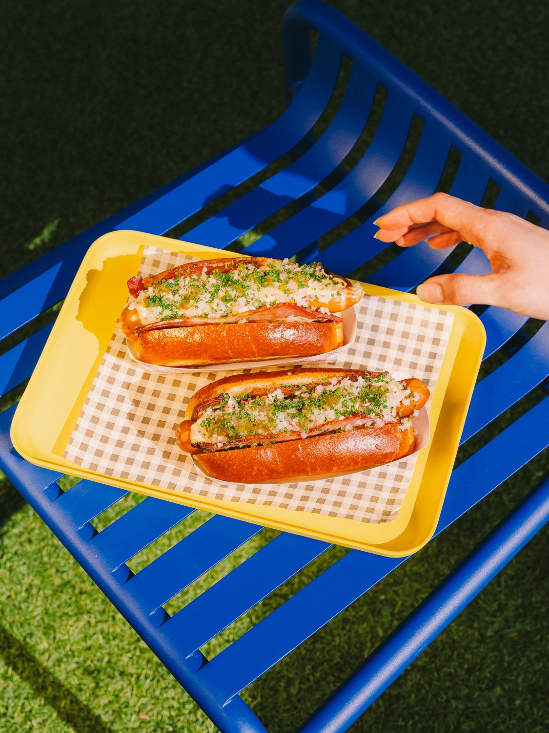 Two lobster roll sandwiches topped with chopped herbs and sauce on a yellow tray, placed on a blue outdoor bench with a grass background.