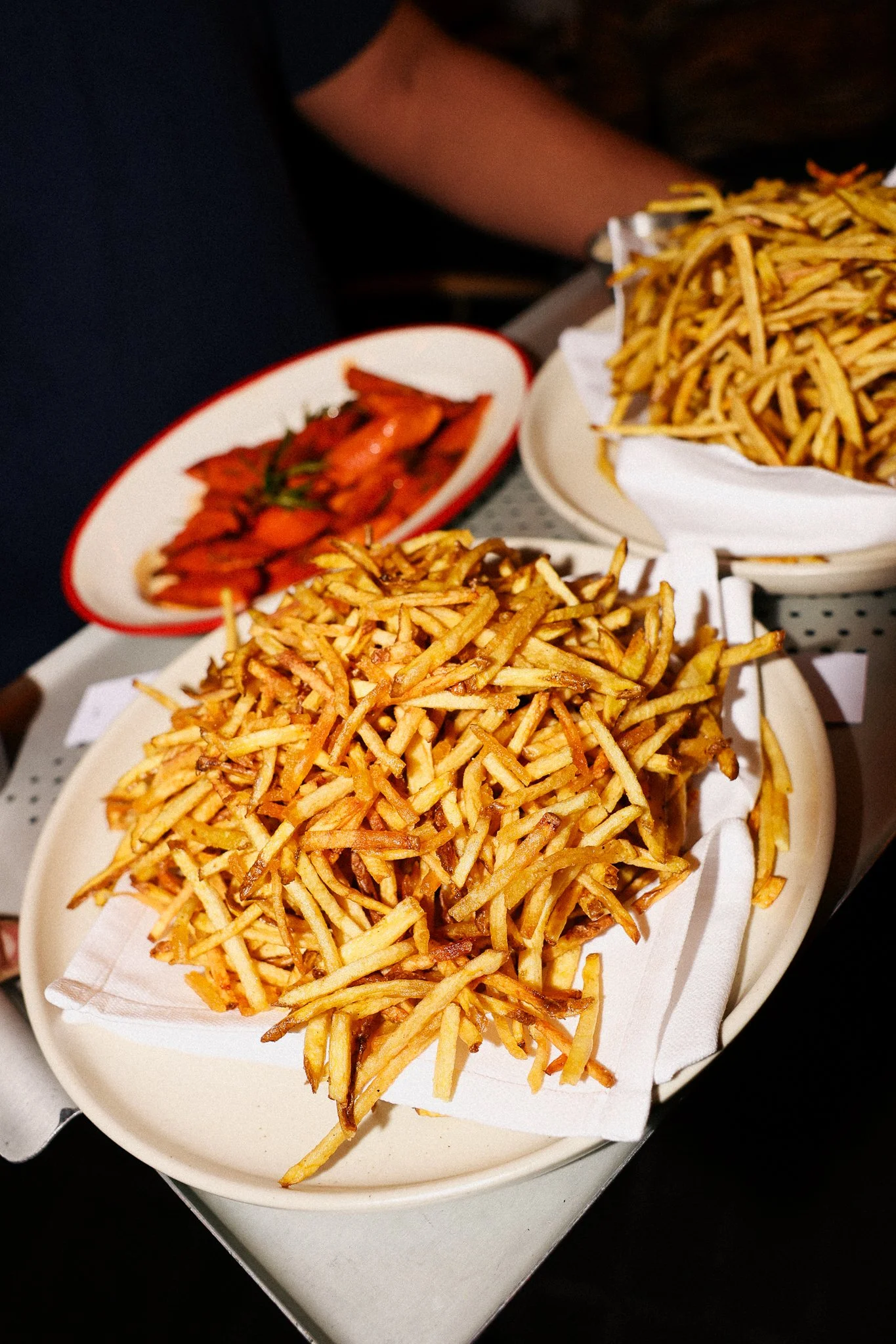 A plate of French fries on a paper-lined basket, with another basket of French fries partially visible, and a small dish of fried shrimp garnished with a sprig of green herbs in the background.