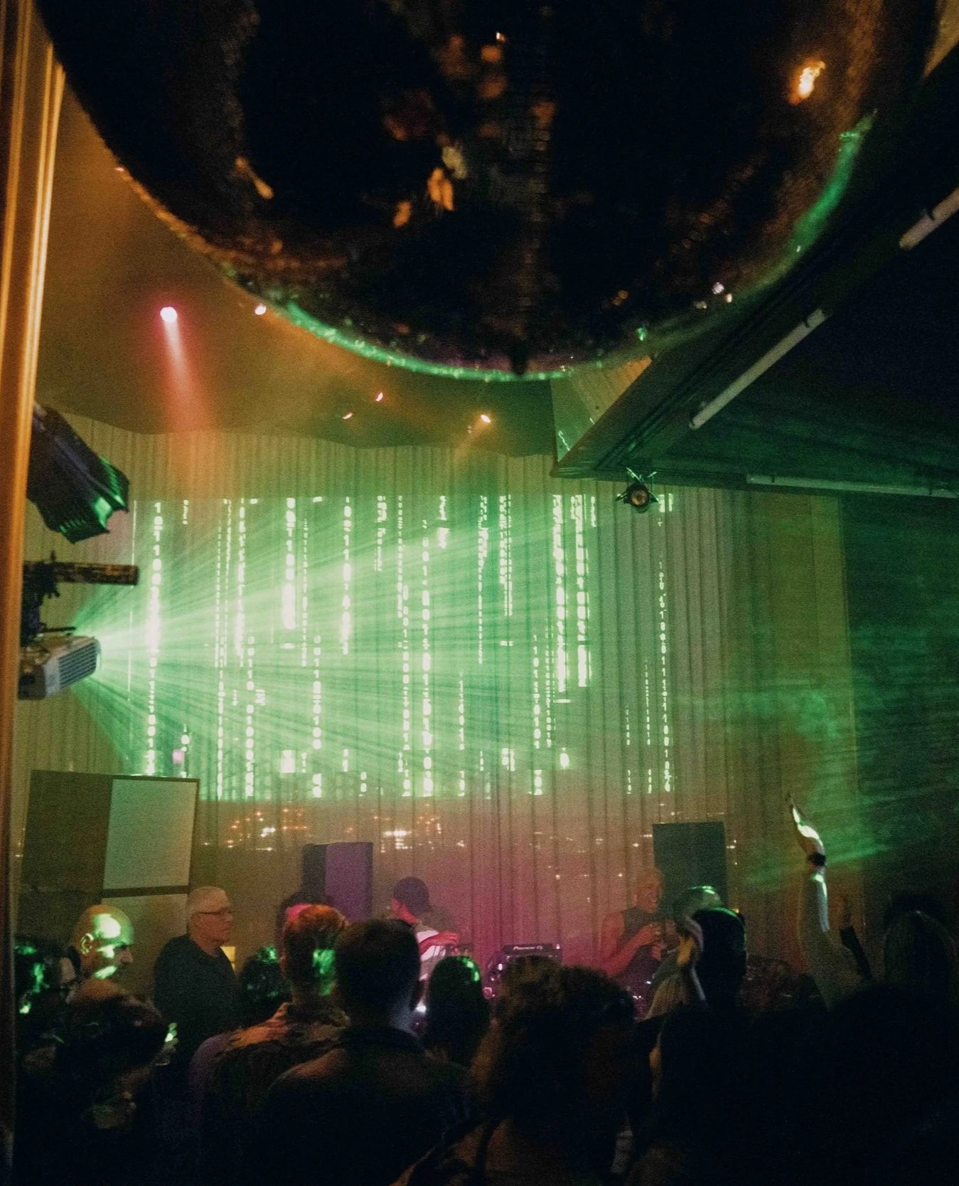 A concert or nightclub scene with people dancing and socializing under green laser lights and a large circular mirror reflecting the scene.