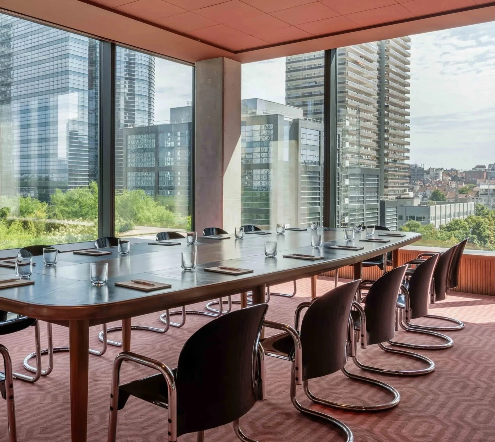 A modern conference room with a long wooden table, black chairs, and glassware set for a meeting. Tall windows overlook a cityscape with high-rise buildings and greenery, and a partially open ceiling reveals an orange-toned ceiling.