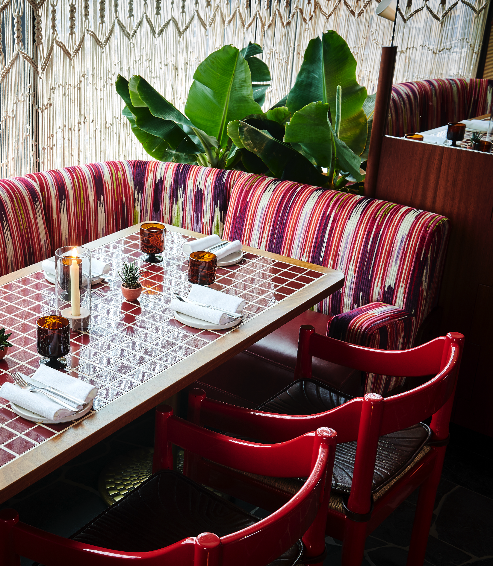 A restaurant dining table set for four with white napkins, utensils, a candle, and a small plant in a pot. The table has a tile surface, and there are chairs with red frames and dark cushioned seats. In the background, there are large green banana plants and a sheer curtain with a beaded design.