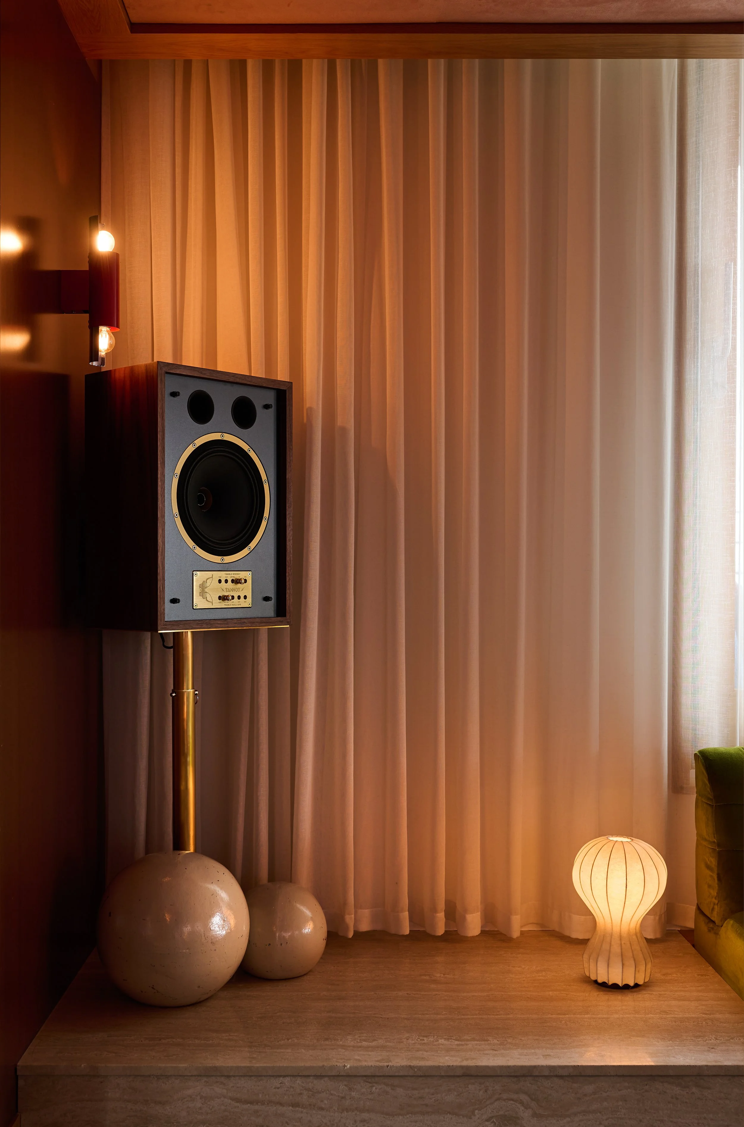 A room corner with beige curtains, a wooden speaker on a stand, three white spherical objects on the floor, a wooden floor lamp, and a yellow velvet chair partially visible on the right.