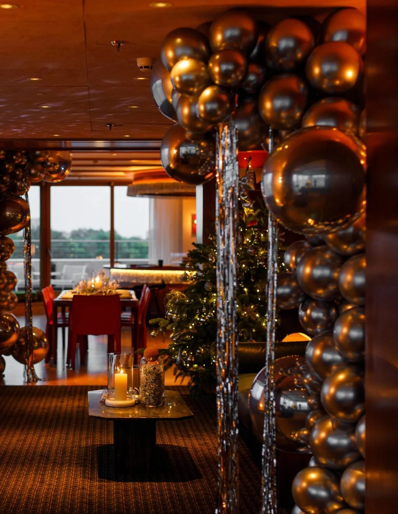 A festive holiday dining area decorated with gold and black balloons, a Christmas tree with lights, and candles on a table.