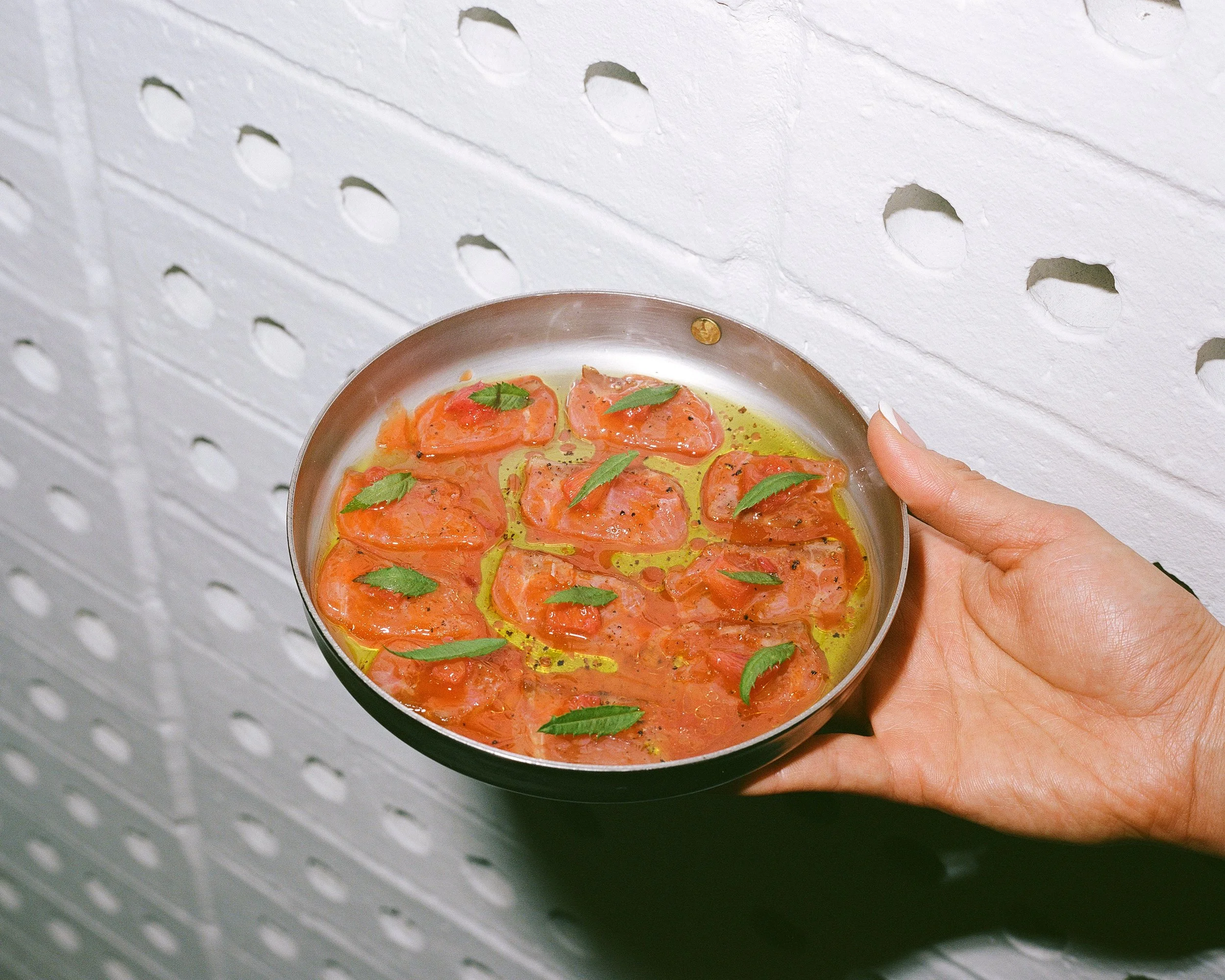 A hand holding a metal plate with sliced salmon topped with herbs and drizzled with oil, against a white textured wall with circular holes.