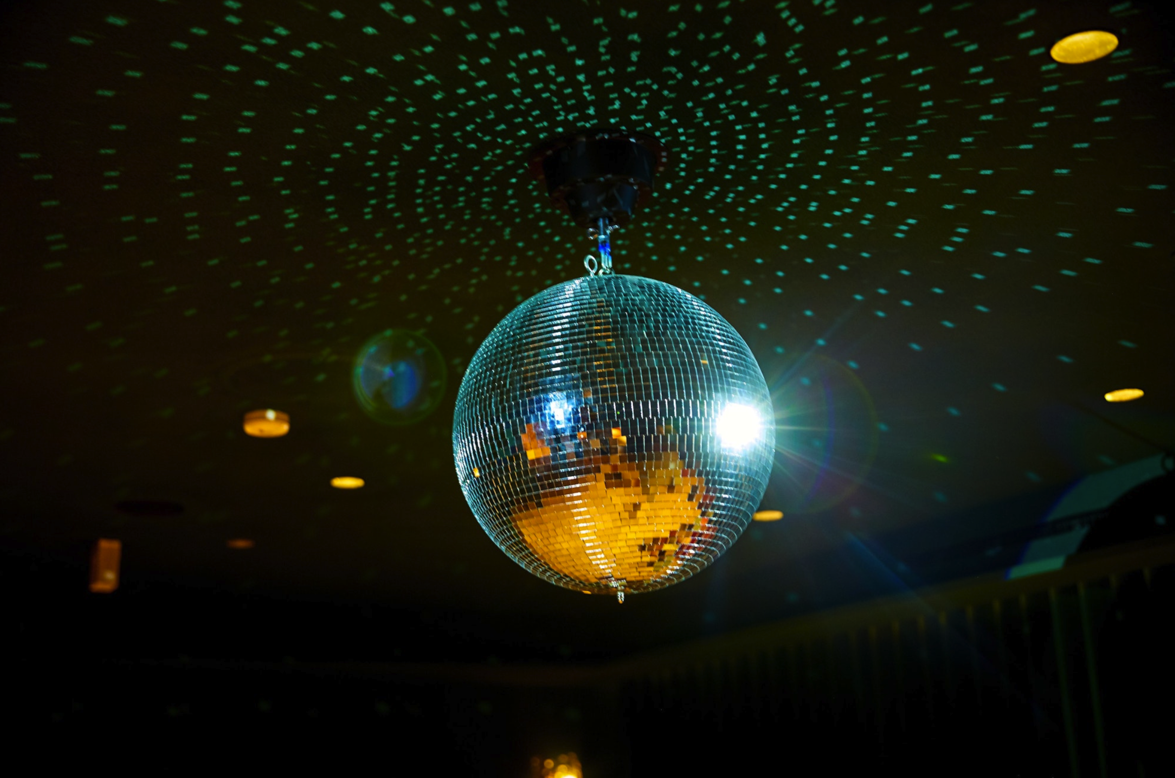 A disco ball hanging from the ceiling reflects light, casting green and gold light spots across the dark room. There are small, circular light fixtures on the ceiling and visible lens flare from the bright reflections.