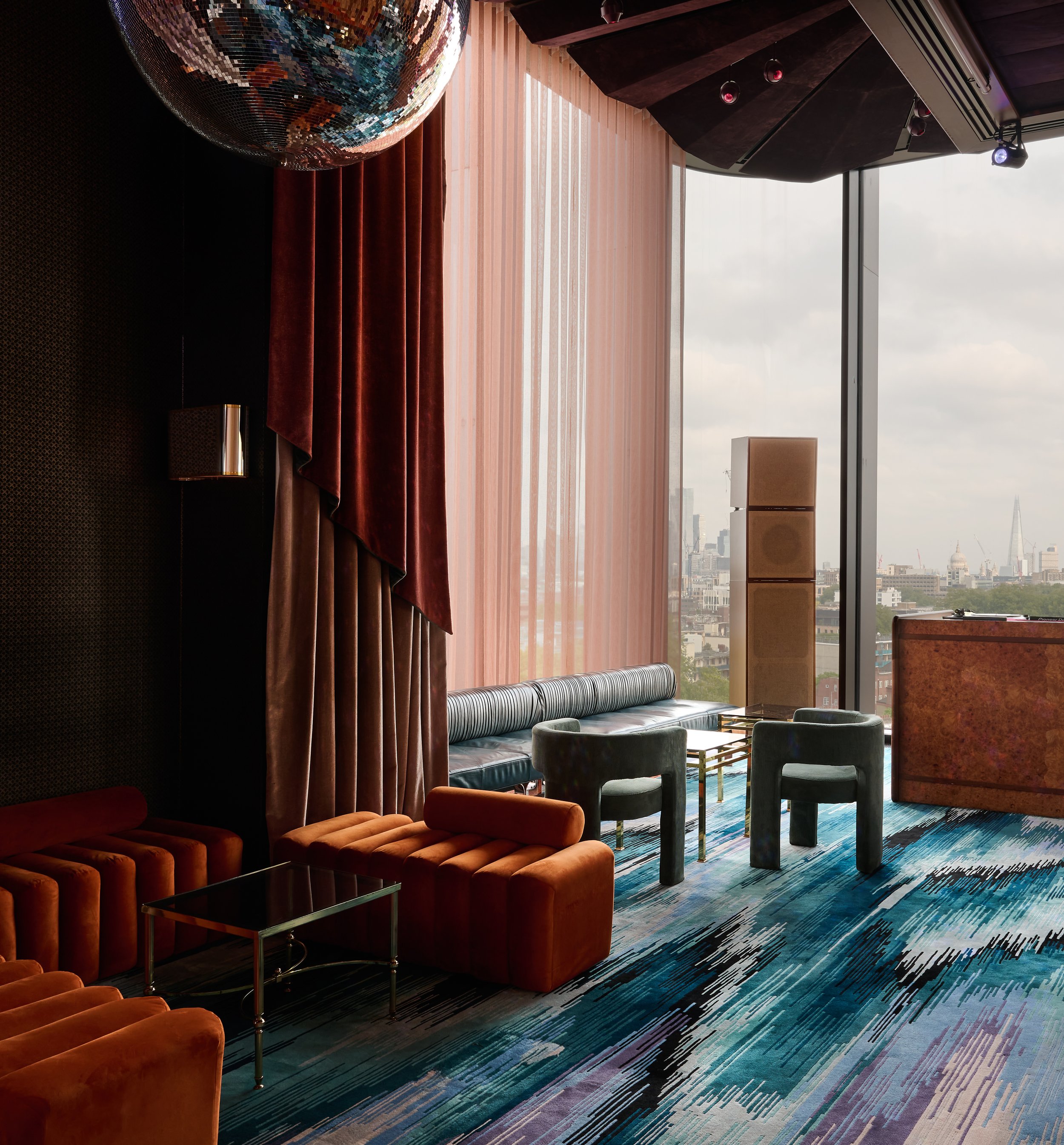 Modern lounge area with orange velvet sofas, green velvet armchairs, a black glass side table, colorful striped carpet, large windows with sheer pink curtains, city skyline view, disco ball on the ceiling, and stage lighting.