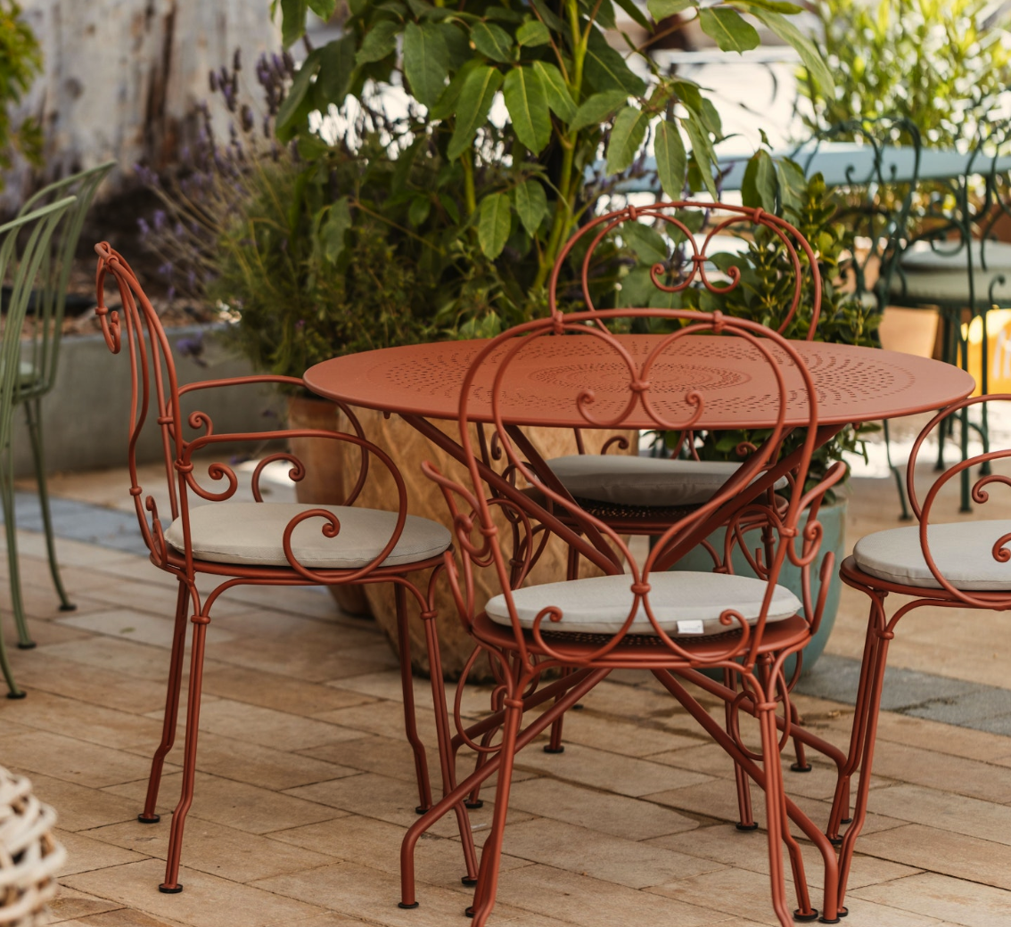 Outdoor patio with a round rust-colored metal table and matching chairs with white cushions, surrounded by large pots of green plants and flowers.